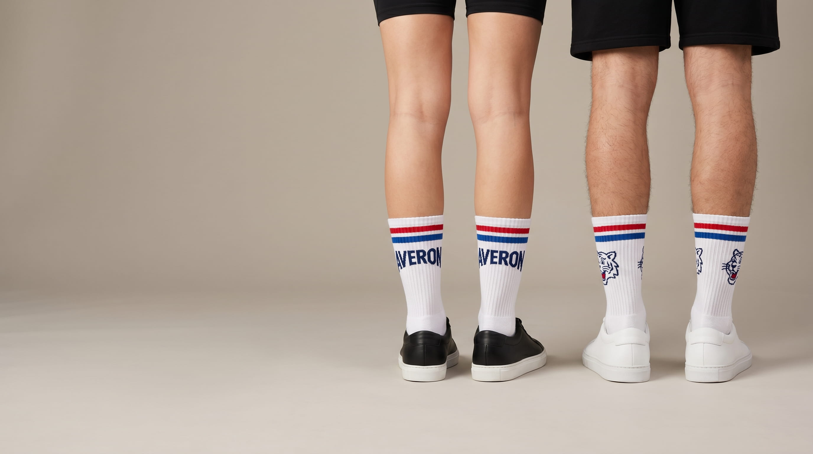 Close-up product-focused shot of two pairs of legs wearing decorated white custom crew socks — one embroidered wordmark, one screen-printed graphic
