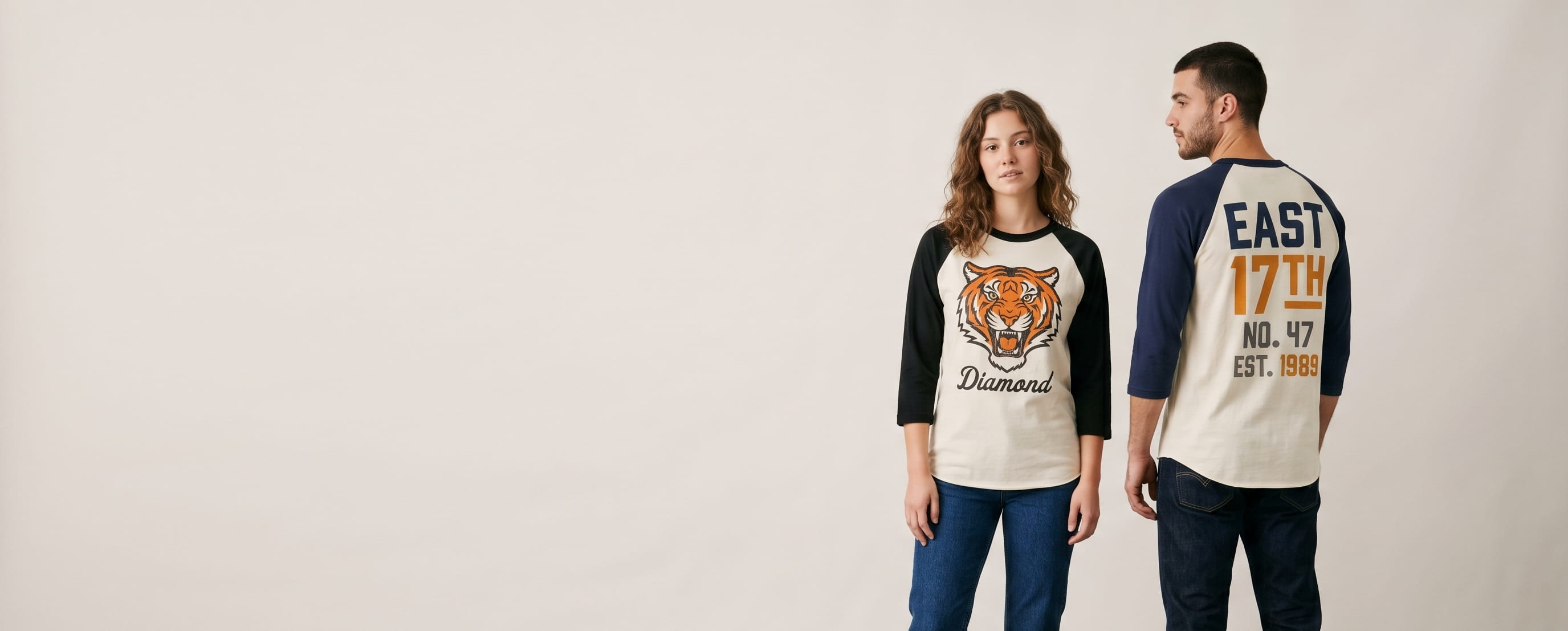 Two lifestyle models in decorated custom 3/4 sleeve baseball raglan t-shirts — woman in cream cotton-poly raglan with classical-statue Medusa screen print + man in cream cotton raglan with bold-typography "EAST 17TH" full-back screen print