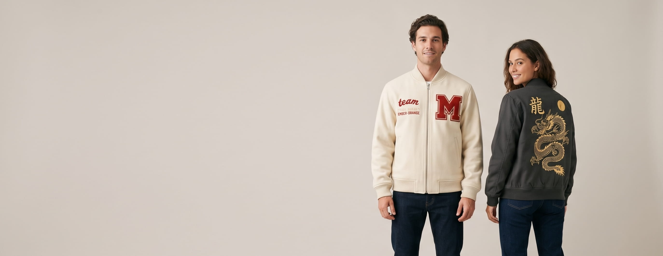 Two lifestyle models in decorated custom bomber jackets — model 1 in cream melton-wool varsity bomber with chenille letter patch and chain-stitch embroidery + model 2 in anthracite cotton-twill bomber with full-back metallic foil decoration