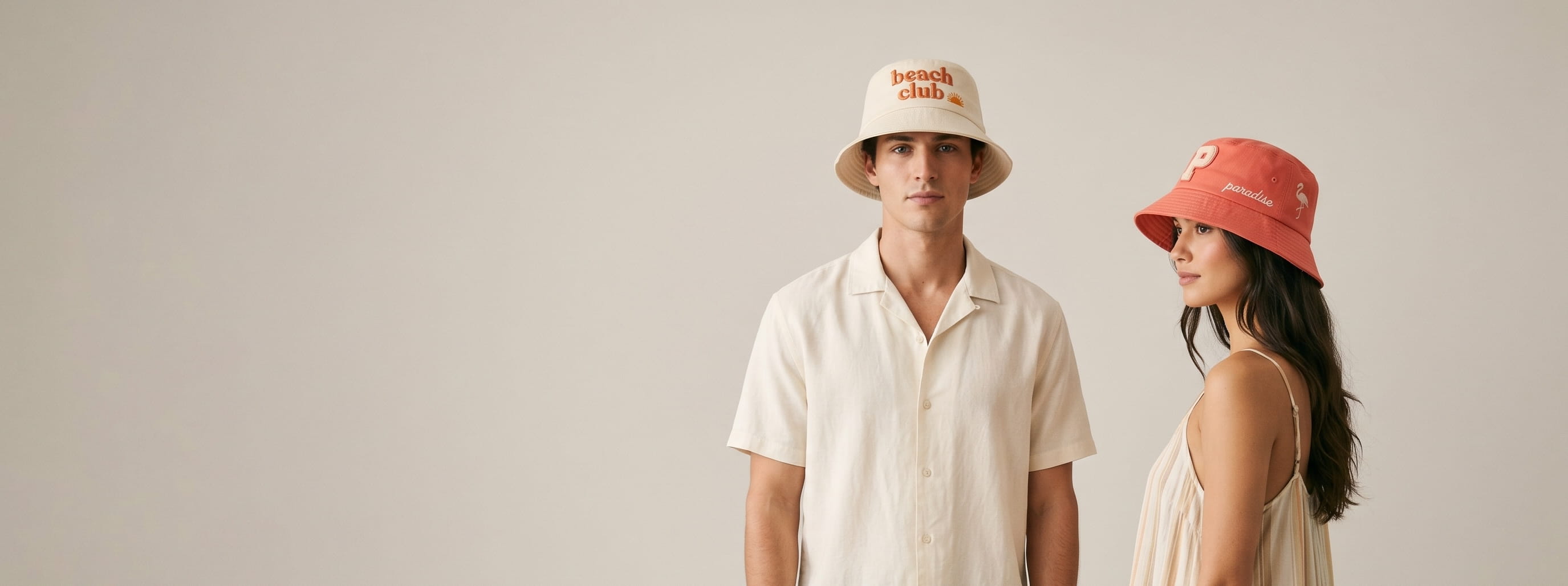 Two lifestyle models in decorated custom bucket hats — man in heritage natural cotton twill bucket hat with 3D puff "beach club" wordmark + tonal sun emblem + woman in saturated coral cotton twill bucket hat with chenille letter patch and chain-stitch "paradise" wordmark