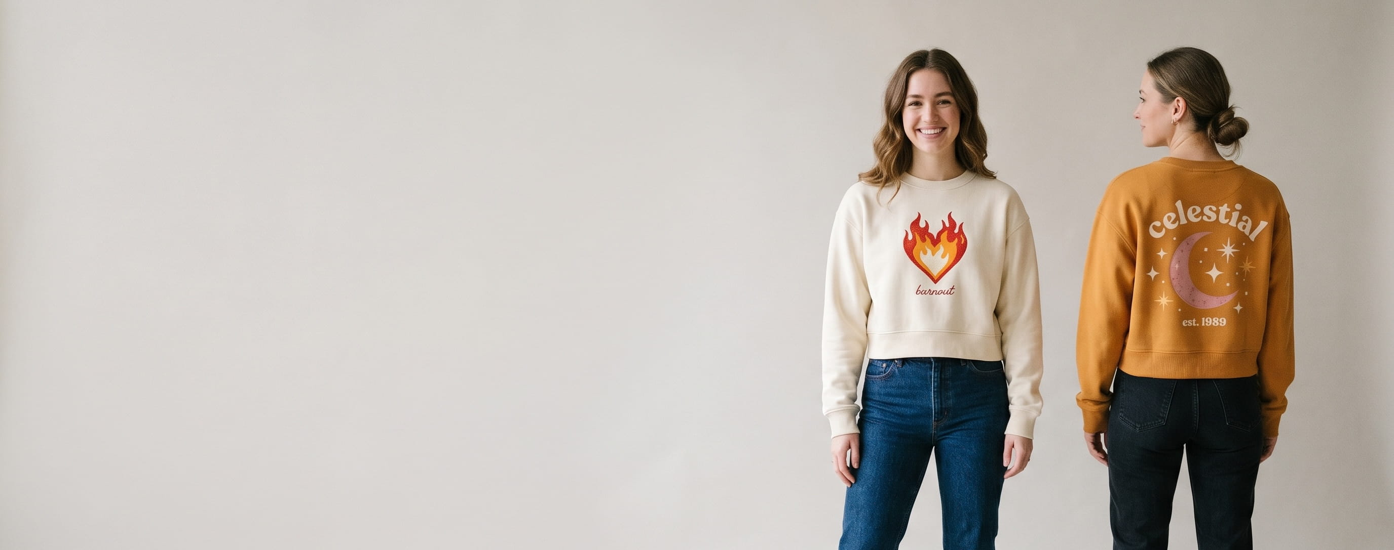 Two women lifestyle models in decorated custom crop top sweaters — model 1 in cream heritage cotton fleece crop crew with hand-illustrated retro-flame-heart "burnout" front-chest transfer + model 2 in warm-amber heritage cotton fleece crop crew with hand-illustrated celestial moon-and-stars "celestial / est. 1989" upper-back composition