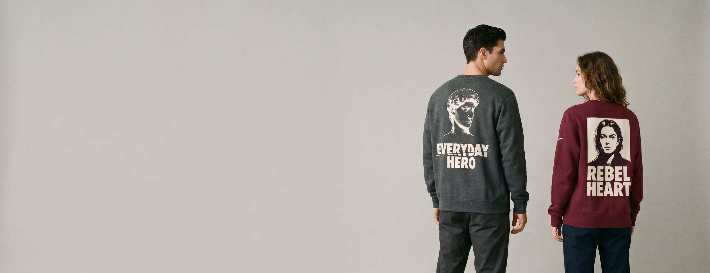 Two lifestyle models in custom Nike sweatshirts — one in a heritage charcoal cotton-polyester brushed-back fleece crewneck sweatshirt with a full-back classical-statue + sliced "EVERYDAY HERO" typography screen print, one in a heritage bordeaux ladies-fit body-conscious cotton-polyester brushed-back fleece crewneck sweatshirt with a full-back photo-portrait + bold "REBEL HEART" typography DTG print — both with the Nike Swoosh at the right chest as on the original Nike blank