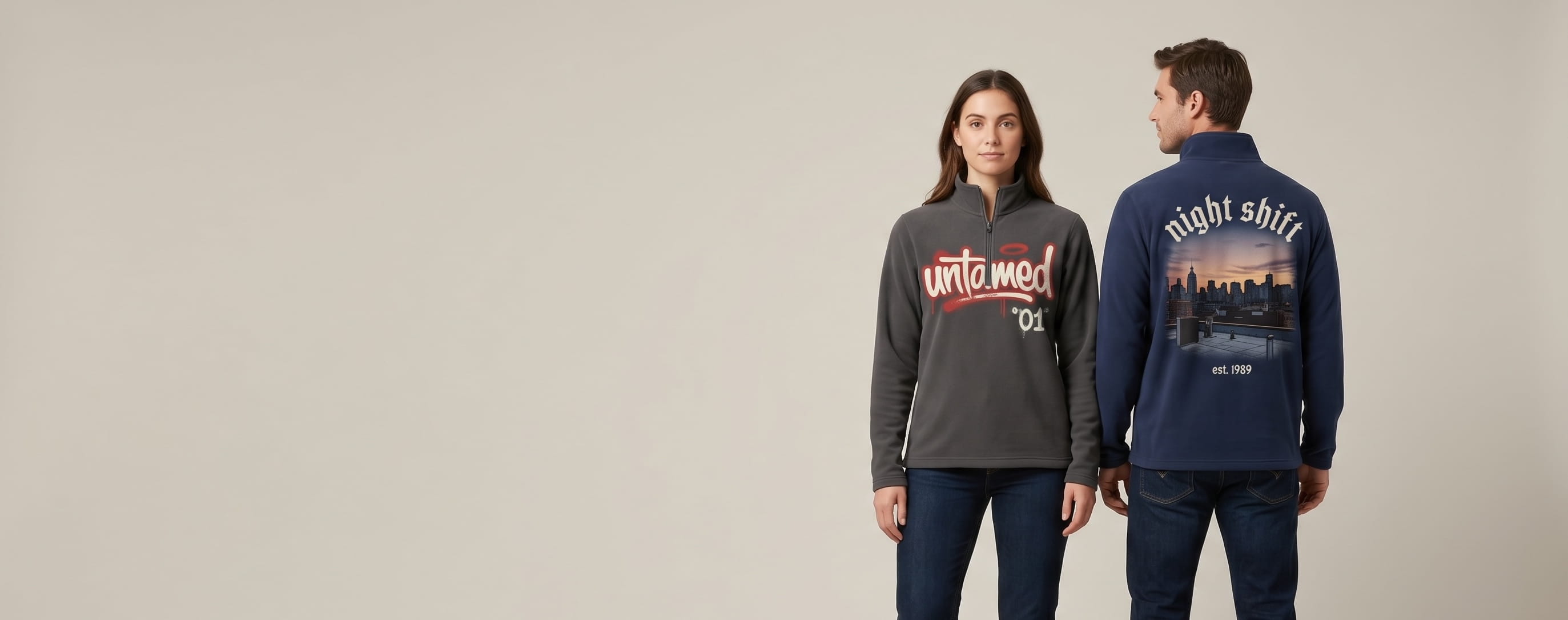 Two lifestyle models in decorated custom half-zip pullovers — woman in anthracite Therma-FIT performance fleece half-zip with hand-illustrated bold graffiti tag "untamed / 01" front-chest transfer + man in navy heritage outdoor fleece half-zip with photo-realistic DTG urban-rooftop-at-dusk scene "night shift / est. 1989" full-back composition