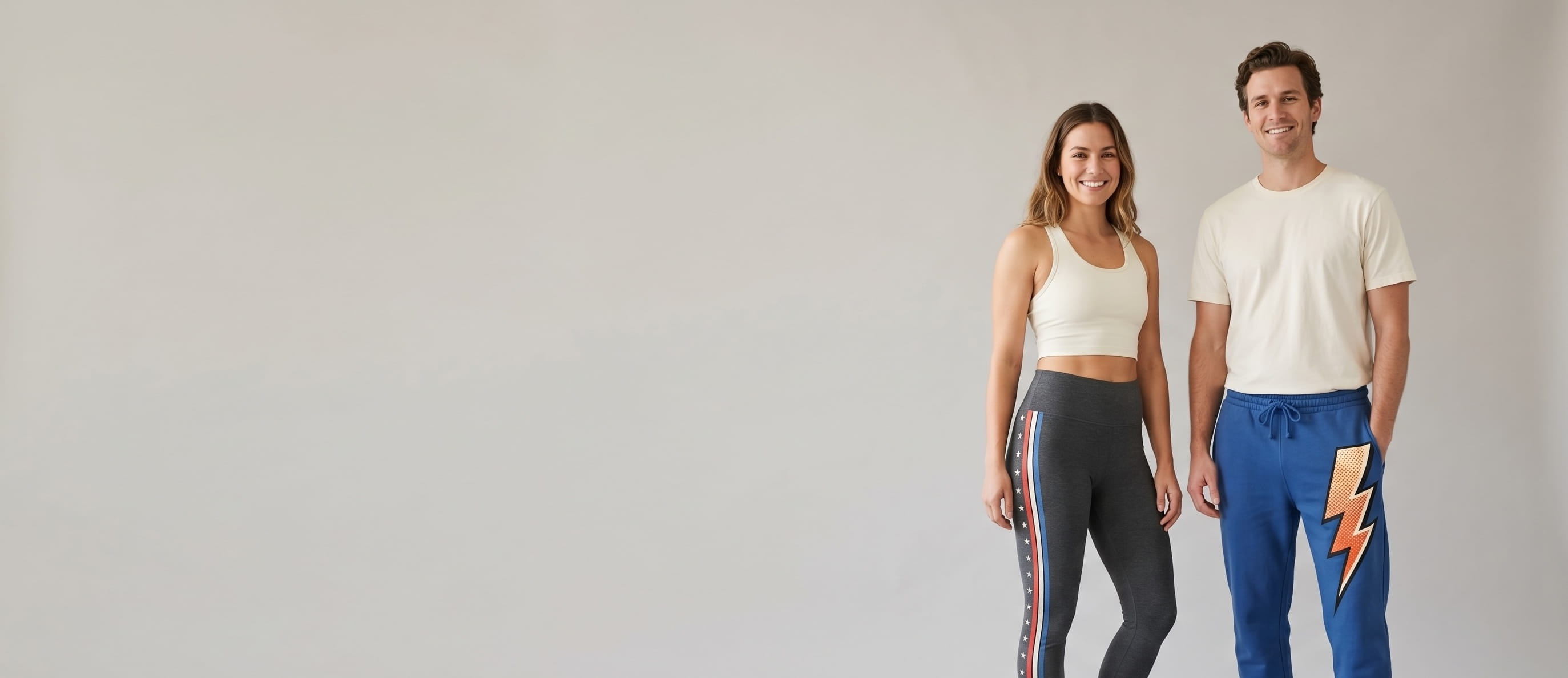 Two lifestyle models in decorated custom bottoms — woman in anthracite heritage cotton-spandex leggings with a full-leg side-seam composition + man in cobalt-blue heritage cotton-fleece sponge fleece sweatpants with a front-thigh full-color DTG composition