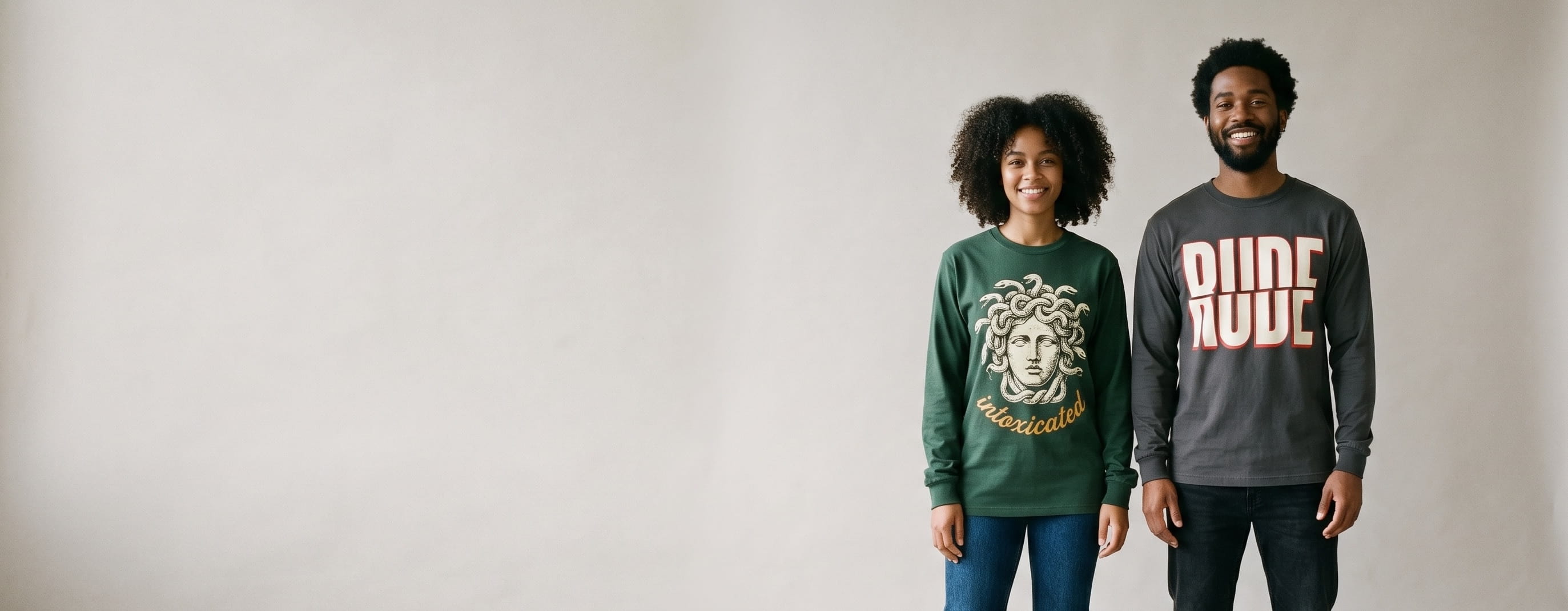 Two lifestyle models in decorated custom long-sleeve t-shirts — woman in heritage forest-green with classical-statue Medusa screen print + man in heritage anthracite with sliced "RUDE" typography screen print