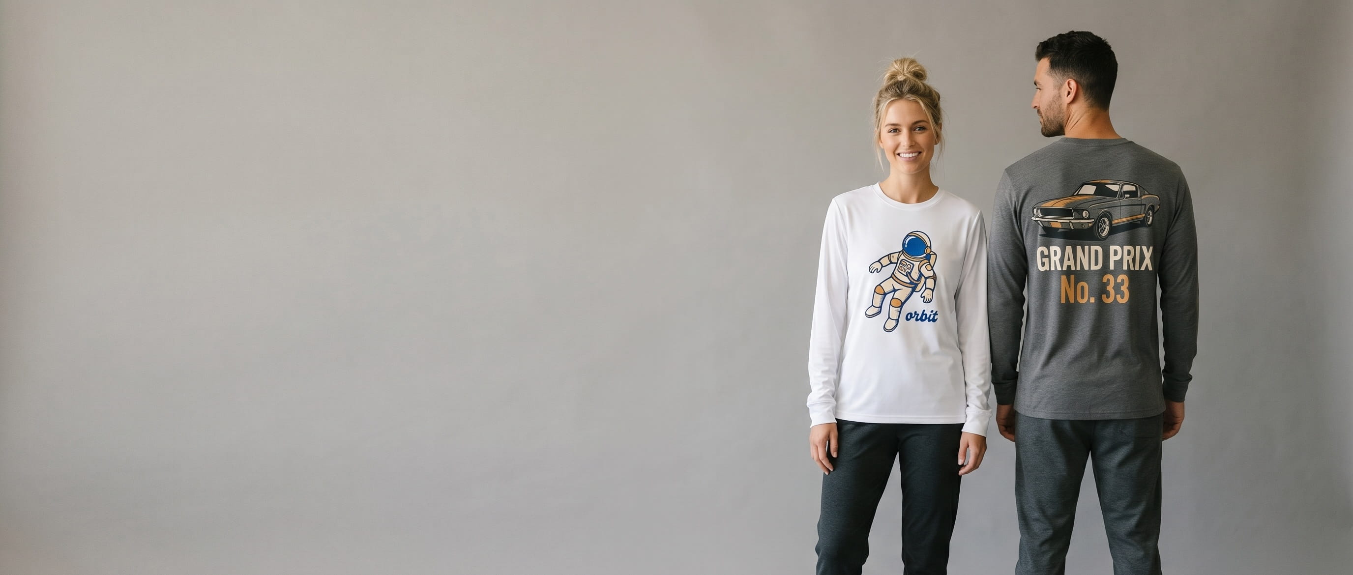 Two lifestyle models in decorated custom performance long-sleeve tees — woman in white performance polyester long-sleeve with cartoon astronaut "orbit" front-chest transfer + man in anthracite performance-blend long-sleeve with muscle-car "grand prix / no. 33" full-back transfer