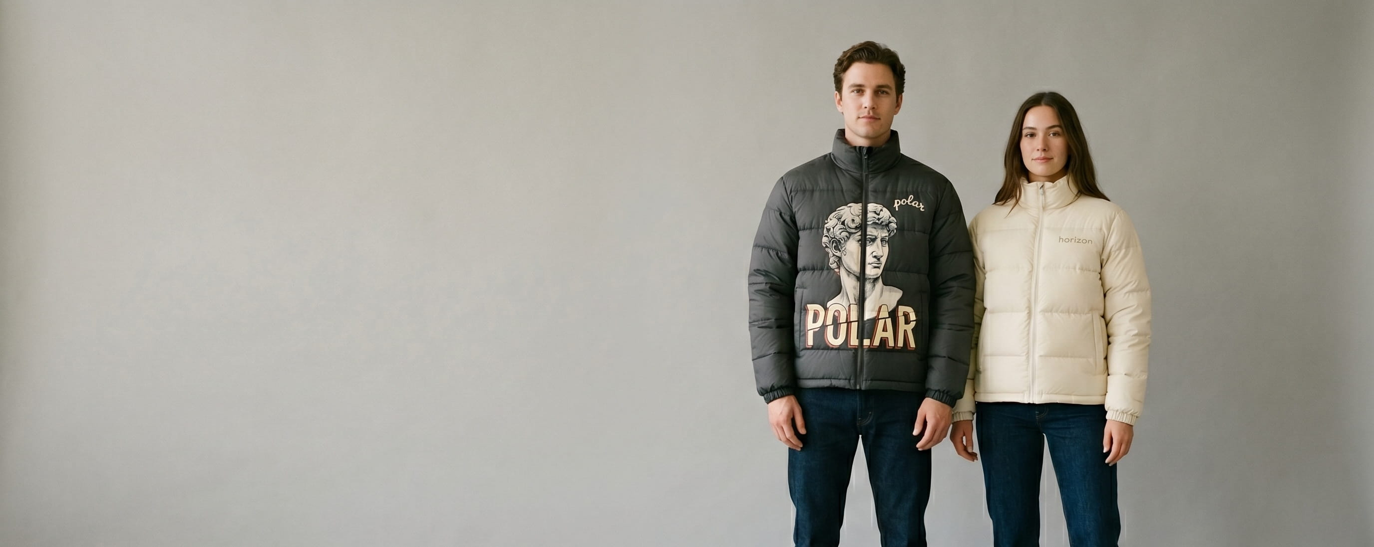 Two lifestyle models in decorated custom puffer jackets — one in anthracite synthetic-insulated puffer with hand-painted artisan transfer + chain-stitch embroidery + one in cream down-insulated puffer with tonal heat-pressed wordmark