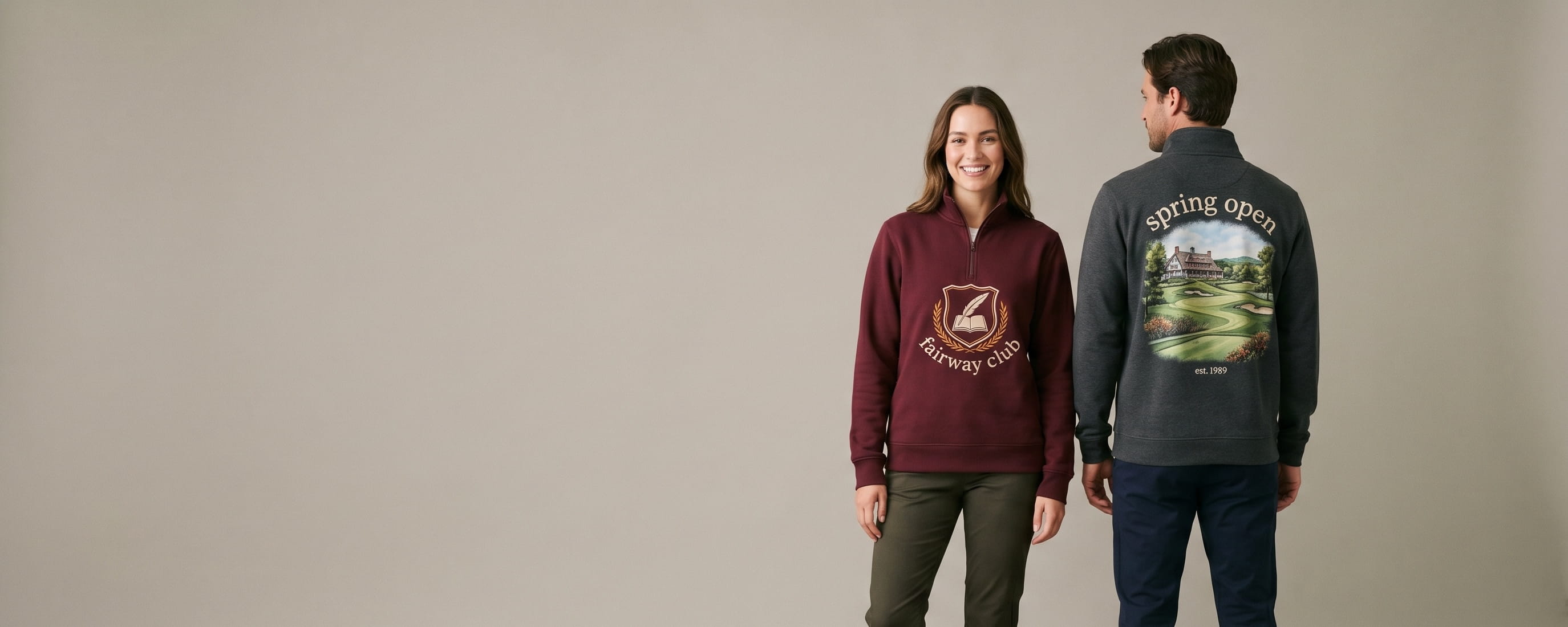 Two lifestyle models in decorated custom quarter-zip pullovers — woman in burgundy heritage fleece quarter-zip with heritage tonal embroidered country-club crest "fairway club" front-chest emblem + man in anthracite performance athletic fleece quarter-zip with photo-realistic DTG golf-course landscape "spring open / est. 1989" full-back composition