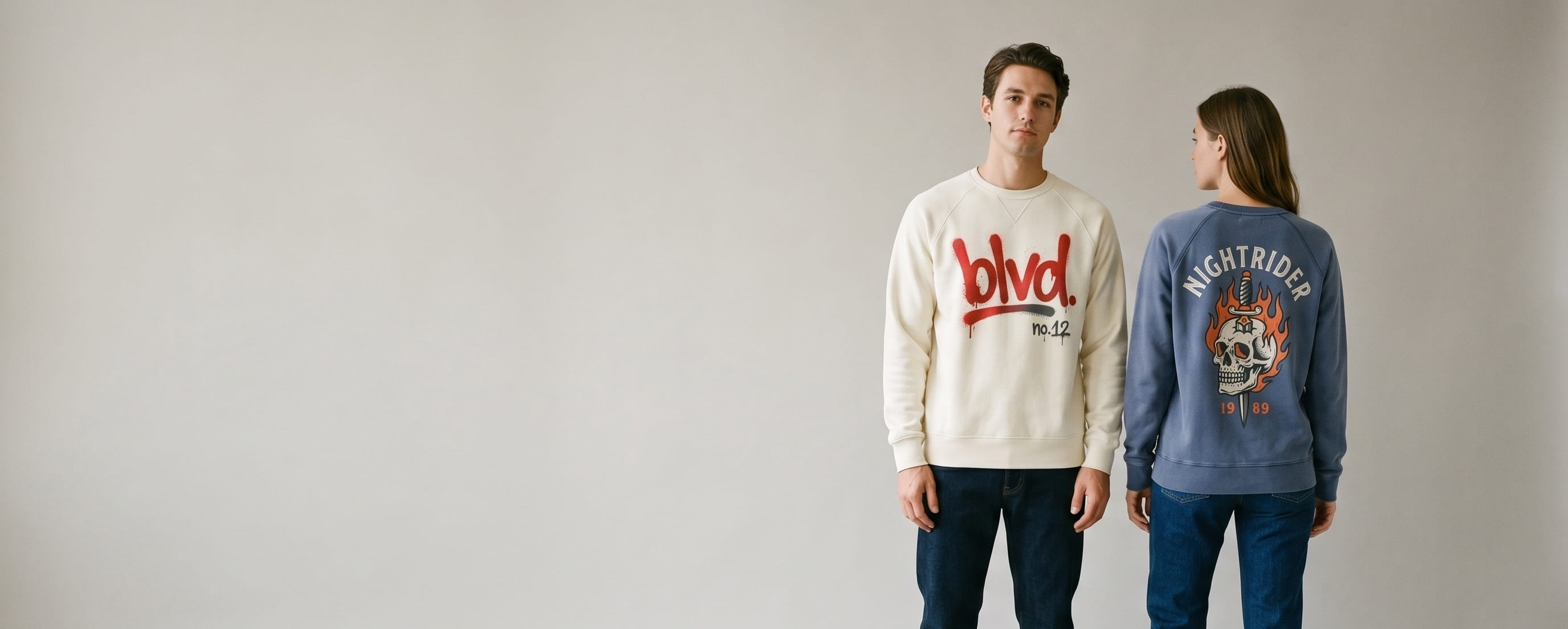 Two men lifestyle models in decorated custom raglan sweaters — model 1 in heritage cream heavyweight cotton fleece raglan with hand-illustrated graffiti spray-paint "raglan / no. 12" plastisol screen print + model 2 in faded indigo garment-dyed cotton raglan with hand-illustrated flaming-skull and dagger plastisol screen-print composition