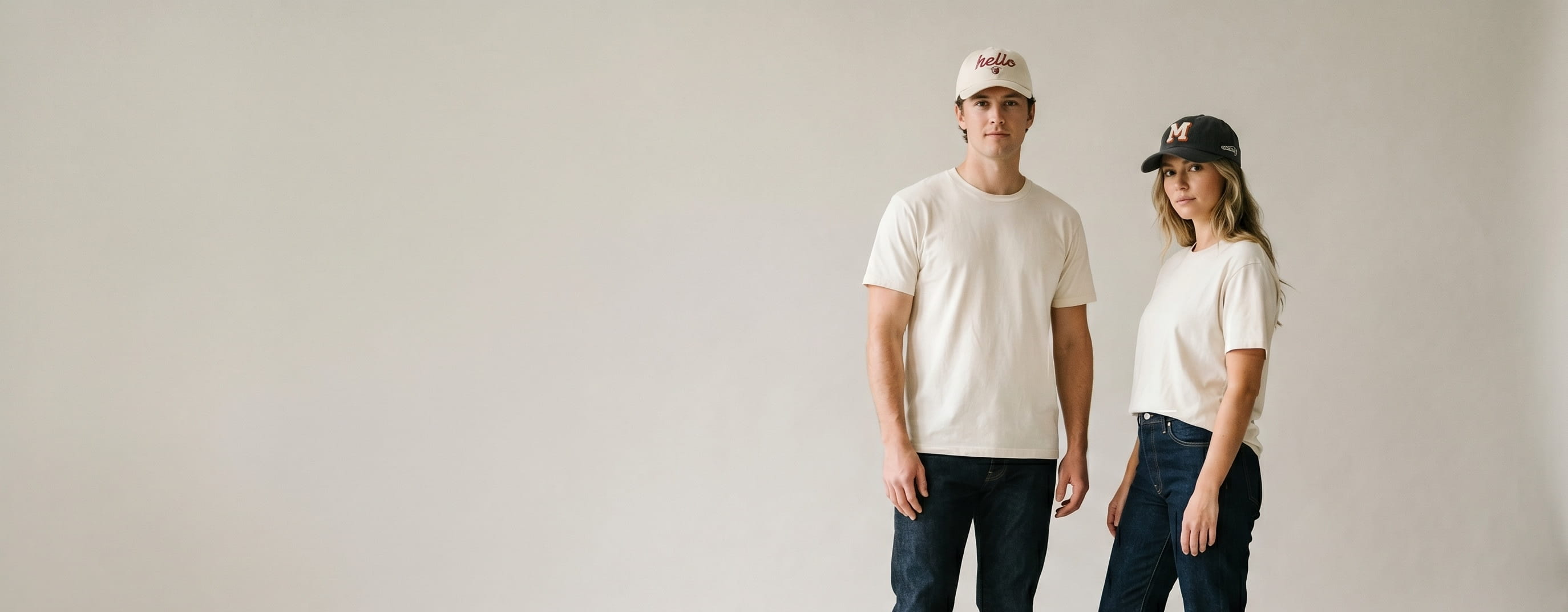 Two lifestyle models in decorated custom six-panel caps — man in heritage natural cotton twill 6-panel cap with heritage flat-embroidered wordmark + tonal shield emblem + woman in heritage charcoal brushed cotton twill 6-panel cap with 3D puff monogram + side panel chain-stitch embroidery