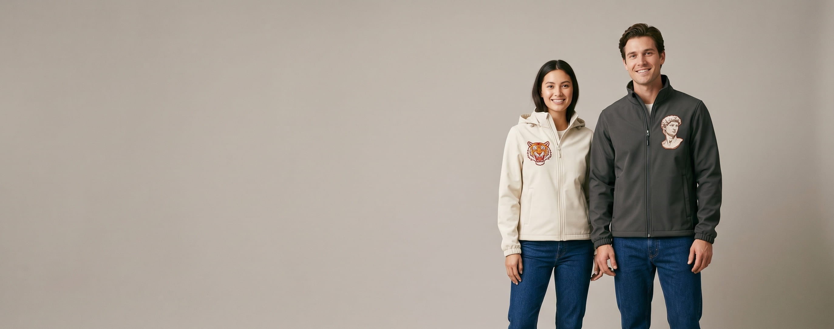Two lifestyle models in decorated custom soft shell jackets — one in anthracite stretch-performance soft-shell with heritage flat-embroidered emblem + one in cream hooded core bonded soft-shell with heritage flat-embroidered illustration