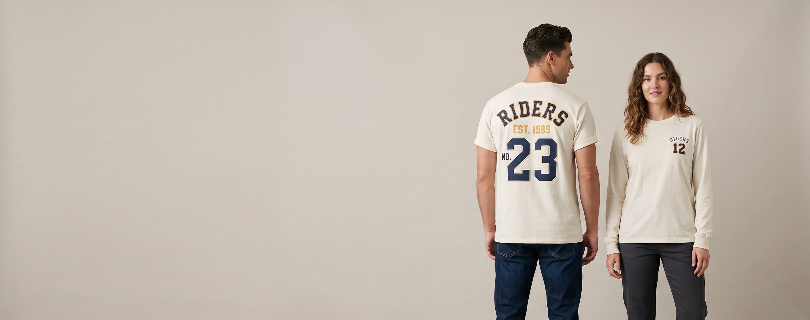 Two lifestyle models in custom team shirts — one in a heritage cream heavyweight cotton jersey tee with a full-back plastisol varsity stacked typography "RIDERS / EST. 1989 / NO. 23"; one in a heritage cream long-sleeve performance tee with a front-chest tonal flat-embroidered "RIDERS" wordmark and small jersey number "12"