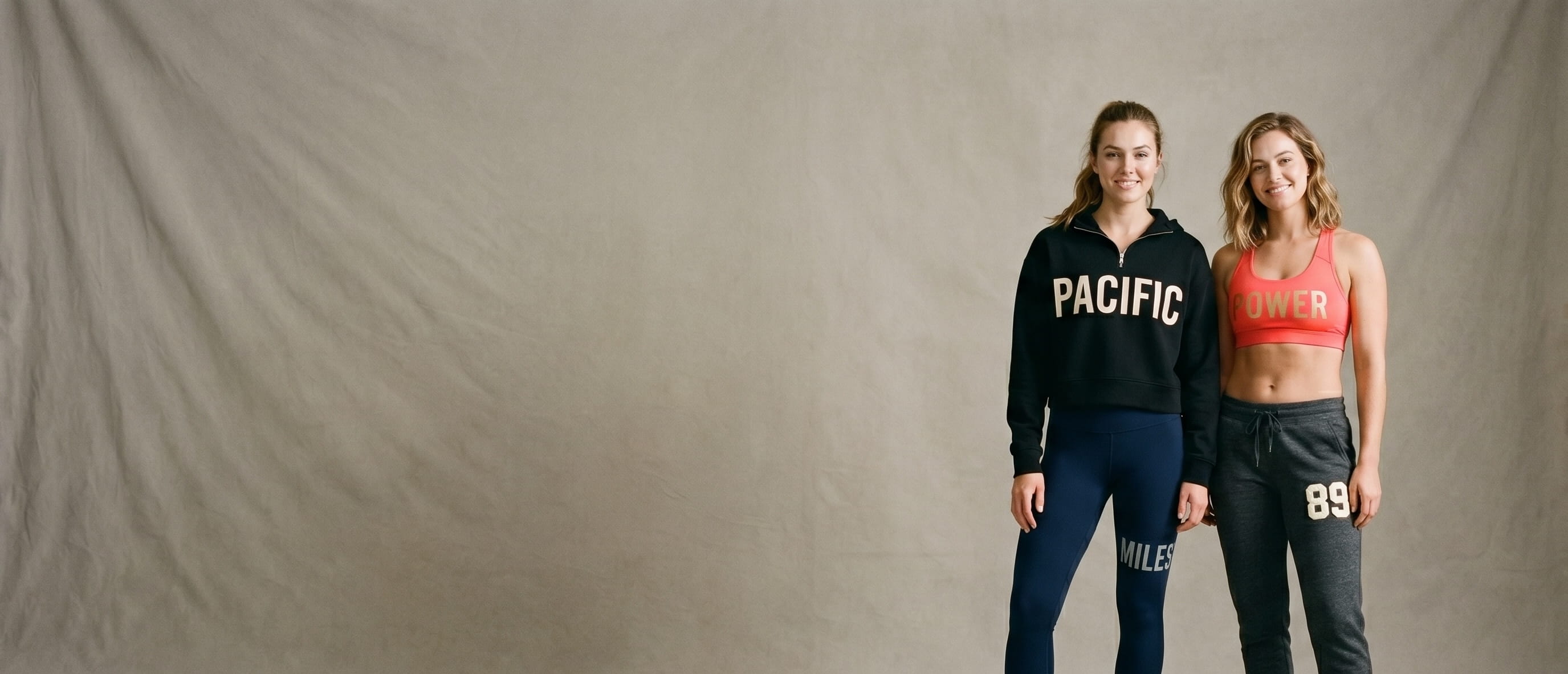 Two fitness-lifestyle models wearing TriDri activewear with bold typography decorations — screen-print, 3M reflective, foil, and puff embroidery on cropped half-zip hoodie, performance leggings, racerback sports bra, and spun-dyed joggers