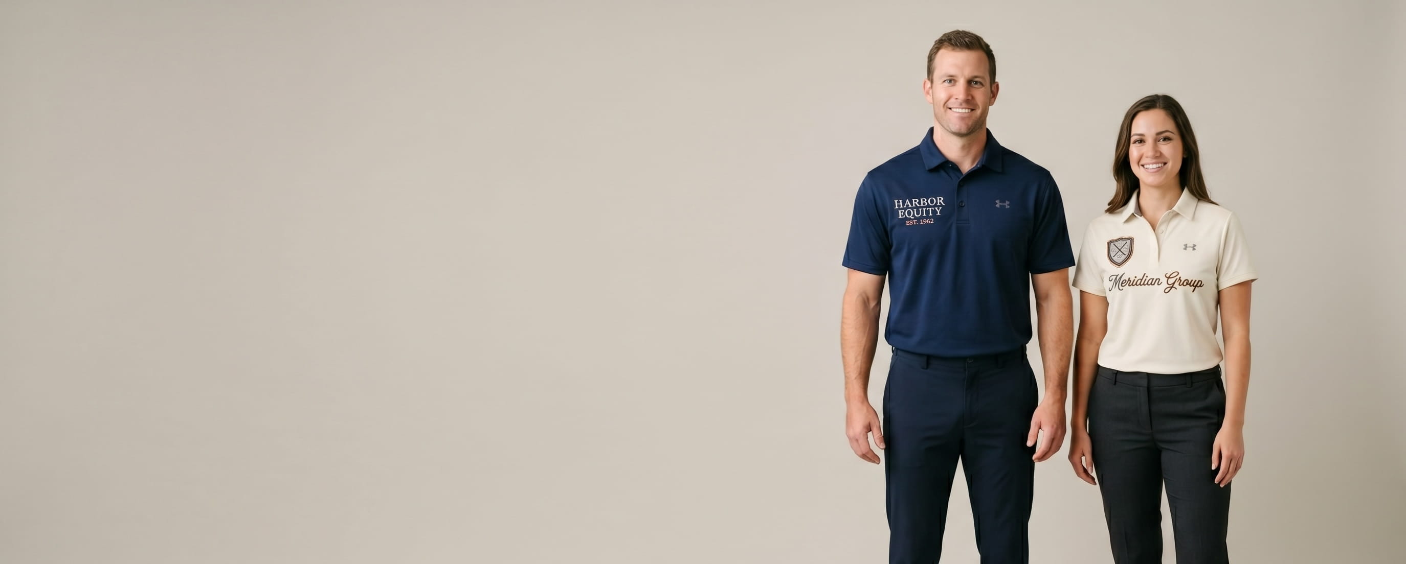 Two lifestyle models in custom Under Armour polos — one in a heritage navy UA Men's Corp Performance Polo with a tonal flat-embroidered "HARBOR EQUITY / EST. 1962" corporate wordmark at front-chest; one in a heritage cream UA Men's Tech Polo with a chain-stitched cursive "Meridian Group" company signature and a small heritage woven crossed-clubs golf-tour shield patch at front-chest