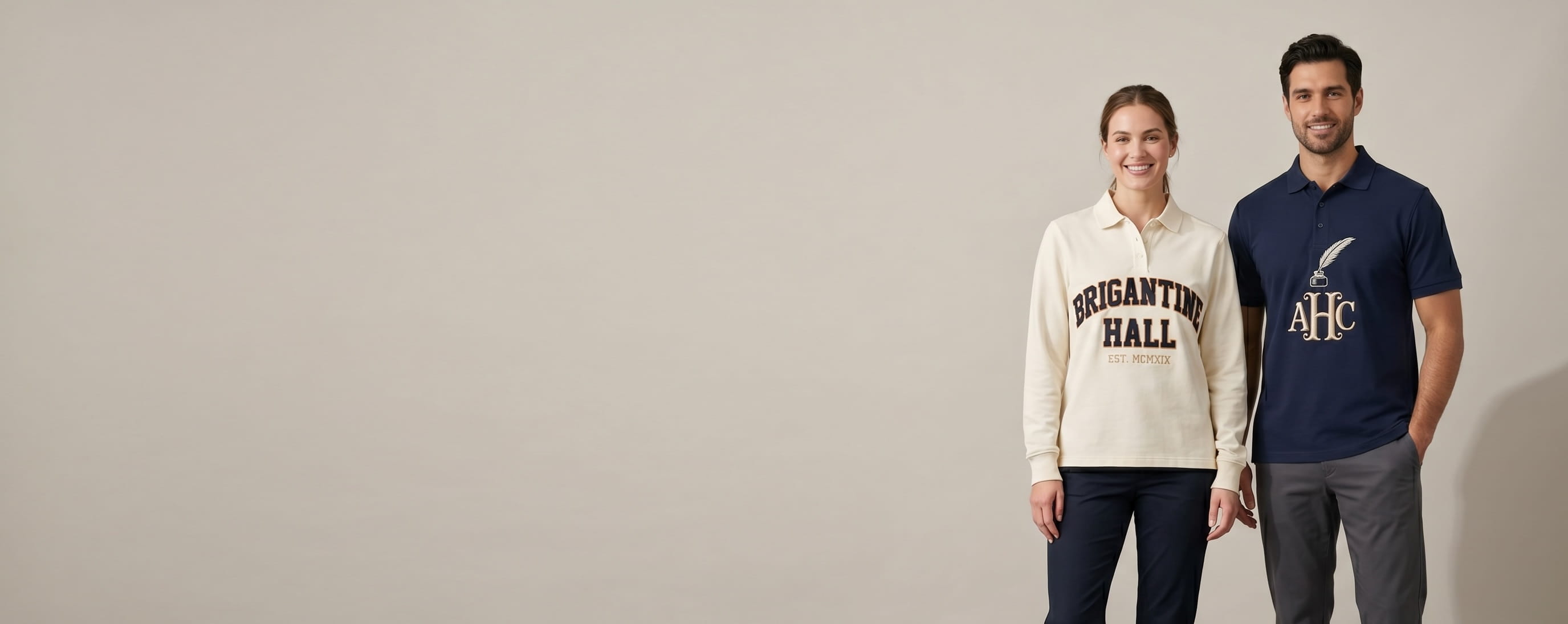 Two lifestyle models in custom embroidered uniform polos — one in a heritage navy short-sleeve performance pique knit polo with a front-chest tonal flat-embroidered intertwined three-letter monogram "AHC" paired with a small hand-drawn quill-and-inkwell silhouette emblem stitched above in cream raised-thread embroidery; one in a heritage cream long-sleeve heritage classic-fit cotton-blend polo with a front-chest 3D puff embroidered bold wordmark "BRIGANTINE HALL" stacked in two rows with a small tonal "EST. MCMXIX" sub-tag centered below in raised foam-backed puff cream-on-cream embroidery