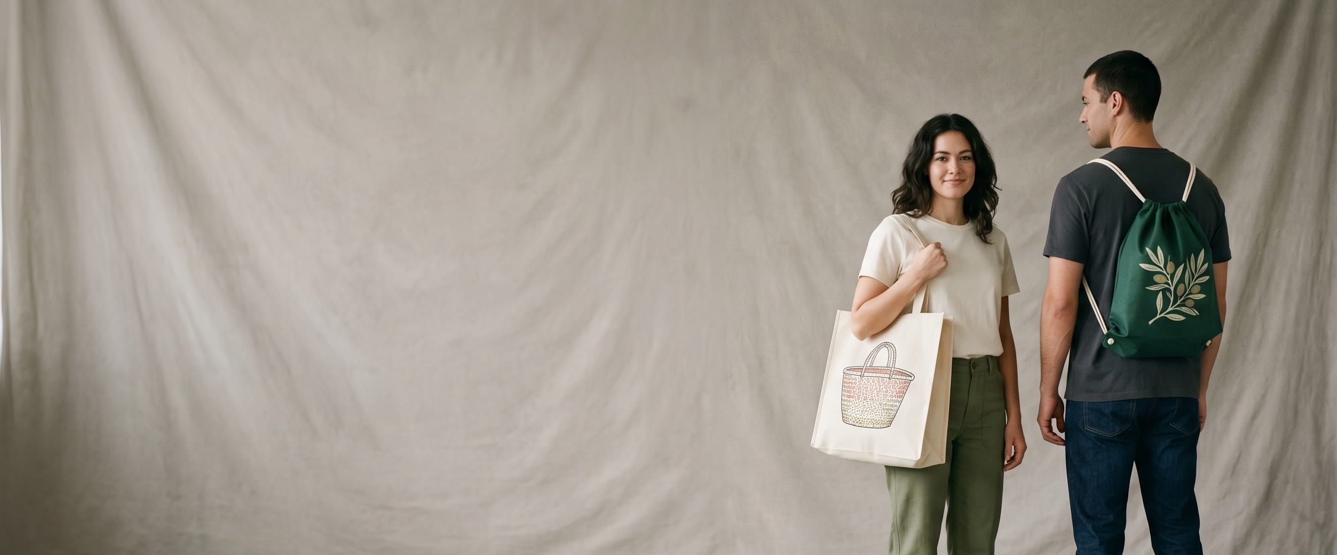 Two community-lifestyle models carrying decorated Valubag pieces — a vintage market-basket plastisol screen-print on a natural-cream non-woven shopping bag and a vintage event-adventure badge heat-pressed transfer on a forest-green non-woven drawstring backpack