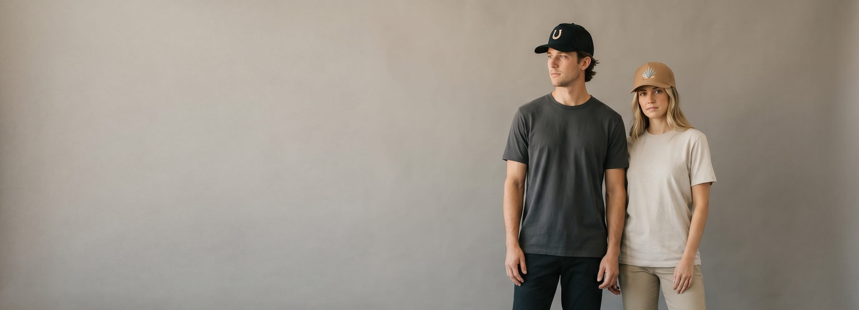 Two casual lifestyle models wearing decorated YP Classics caps — vintage horseshoe embroidery on the man's deep-black 100% cotton retro trucker and vintage agave-plant embroidery on the woman's kraft-tan poly-spandex perforated snapback