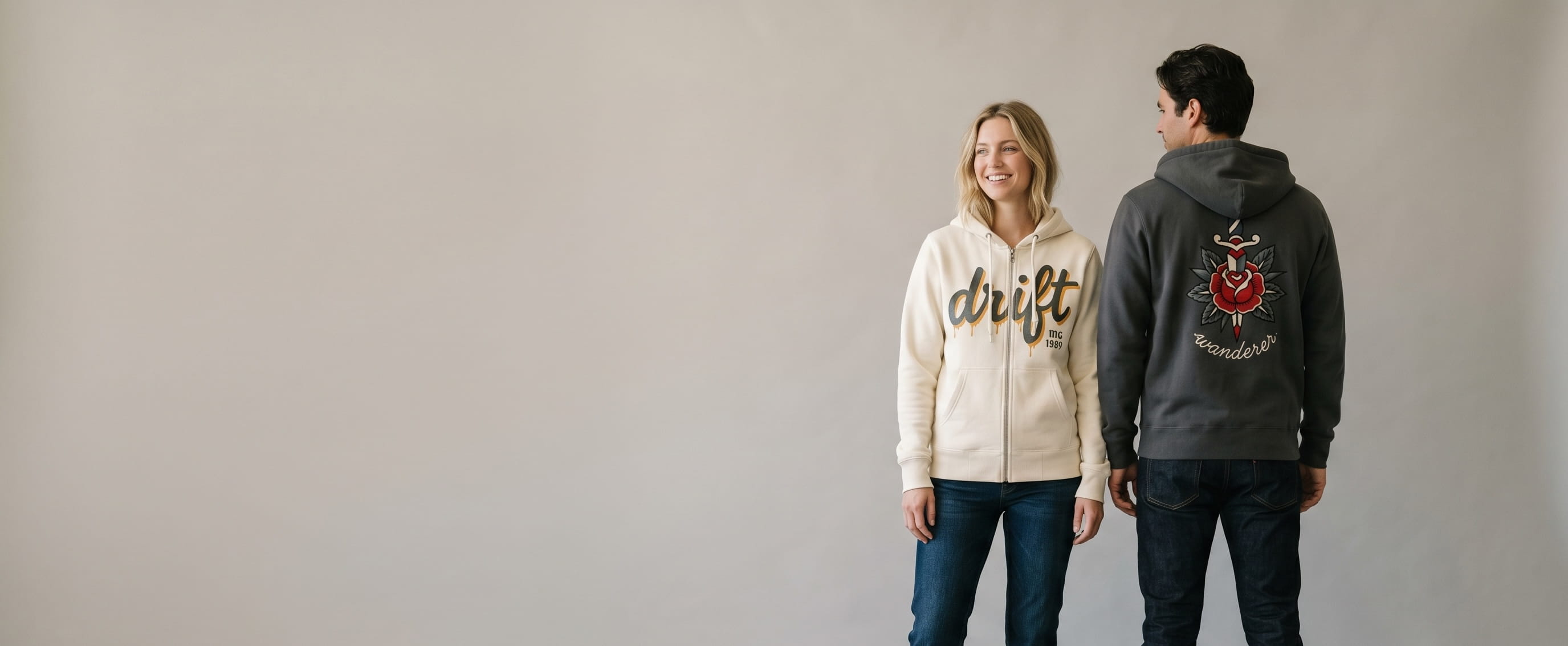 Two lifestyle models in decorated custom full-zip hoodies — woman in cream heavyweight cotton fleece full-zip with handpainted drippy "drift" front-chest transfer + man in anthracite heavyweight cotton fleece full-zip with embroidered tattoo-flash rose-and-dagger + "wanderer" cursive script full-back transfer
