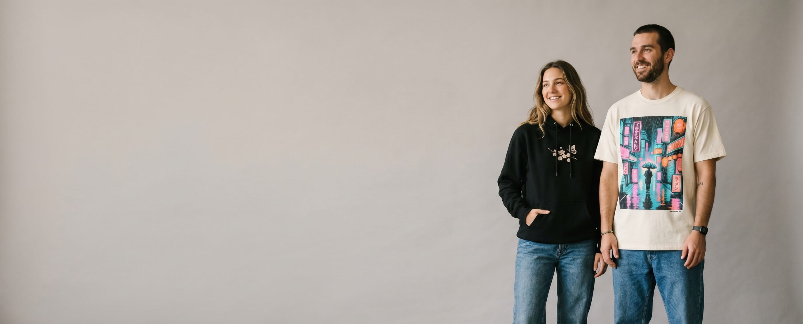Two streetwear-lifestyle models wearing decorated American Apparel — Tokyo neon street-scene DTG on the man's heritage cream heavyweight cotton tee and hand-sketched cherry-blossom + butterfly embroidery on the woman's deep-black ReFlex Fleece hoodie