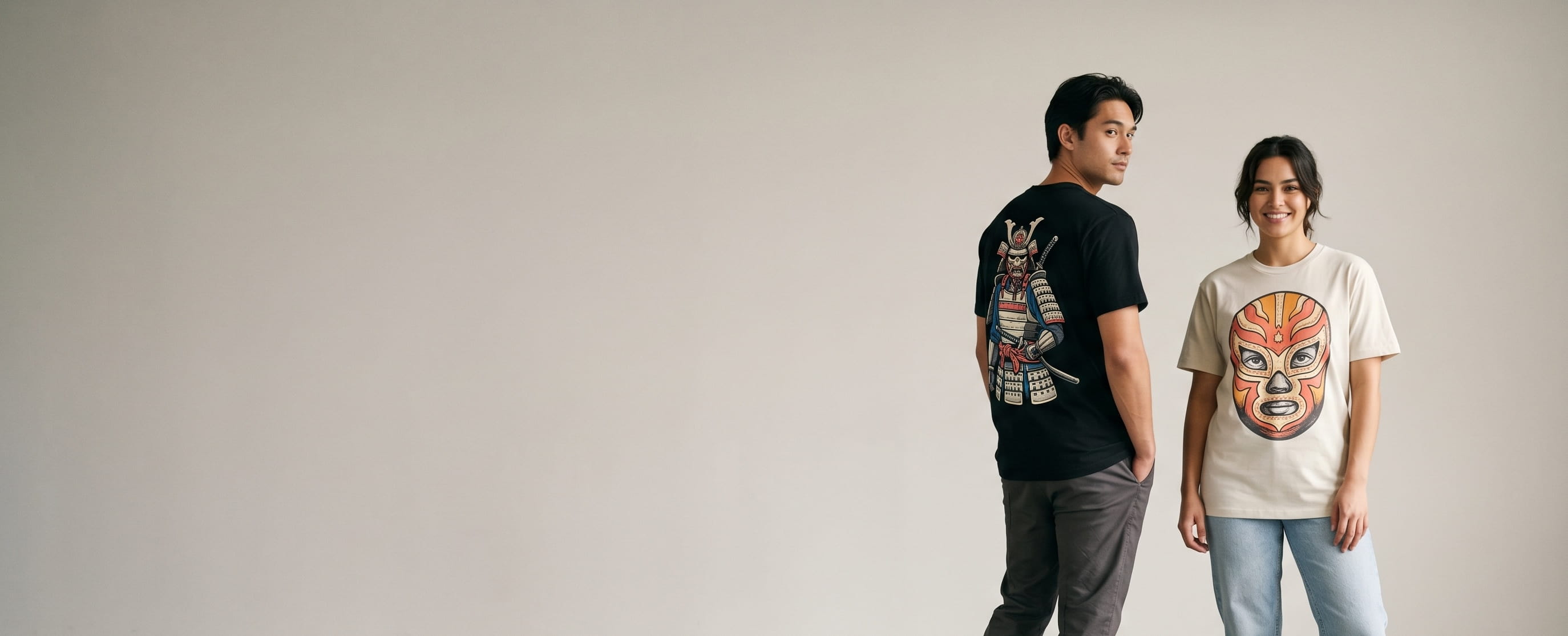 Two streetwear lifestyle models wearing decorated AS Colour — a full-back cyber-samurai plastisol screen print on the man's jet-black heavy tee and a full-chest lucha-libre-mask full-color DTG on the woman's bone Staple tee