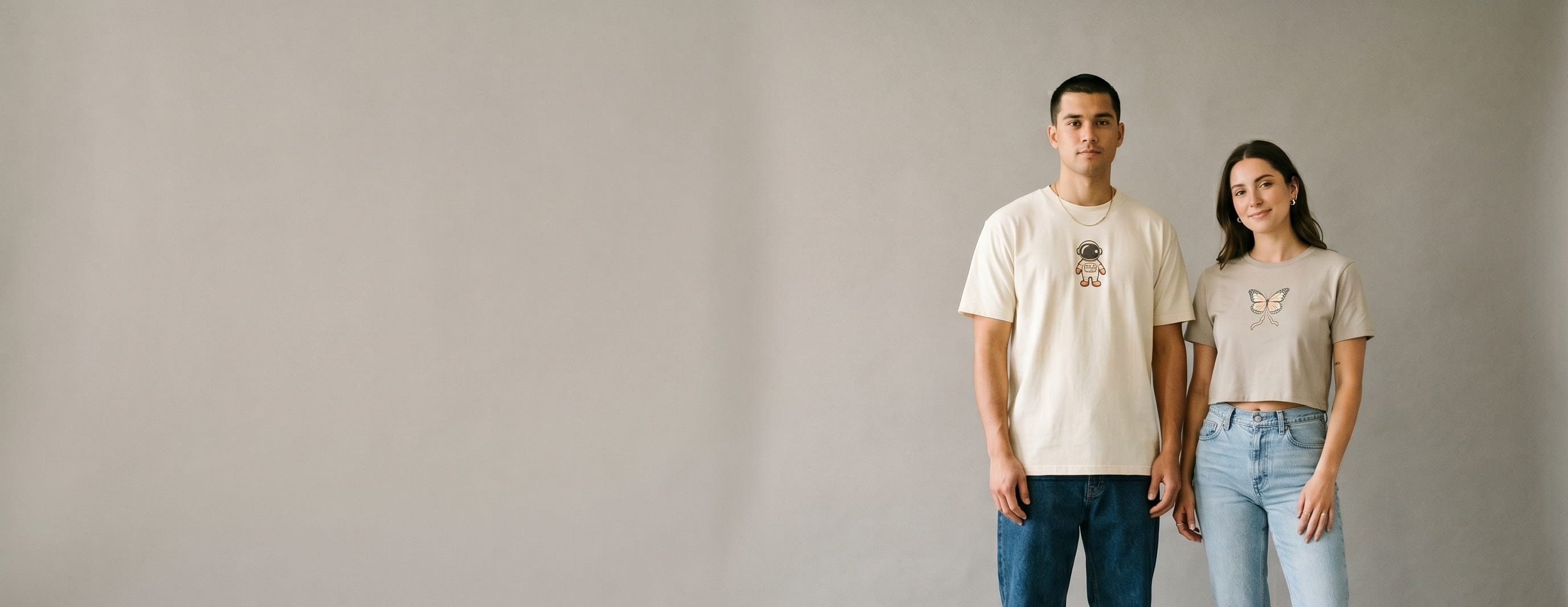 Two streetwear-capsule lifestyle models wearing decorated Bella + Canvas — Pinterest 2026 cartoon astronaut emblem embroidery on the man's heritage cream Heavyweight Street tee and DBTK butterfly + script heat-press transfer on the woman's heritage stone Women's Crop Tee