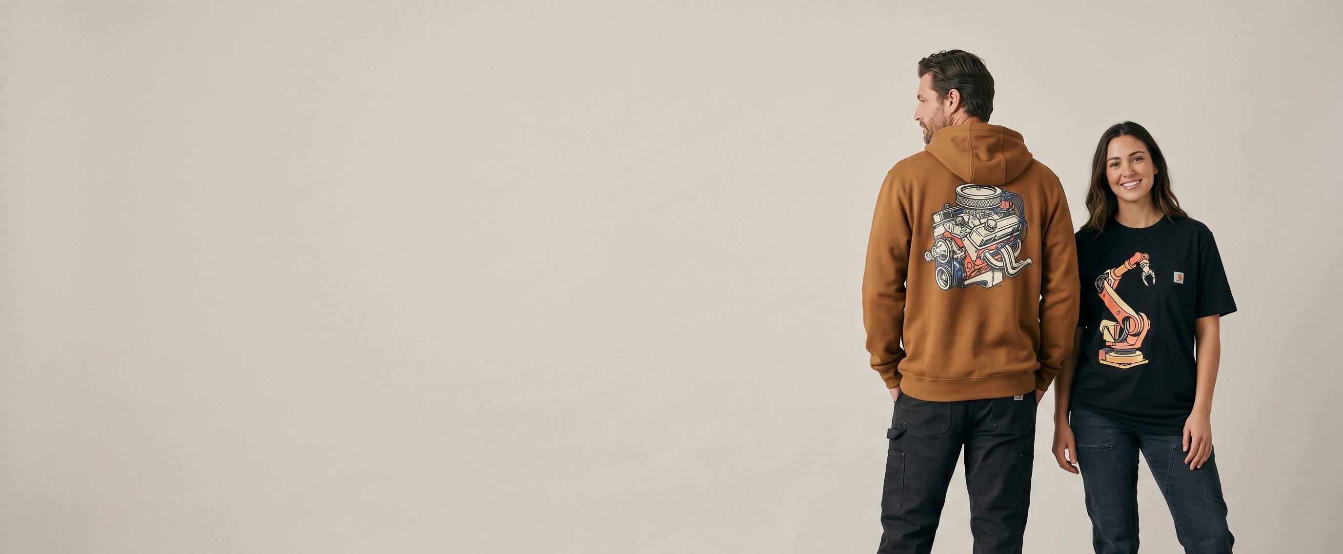 Two workwear lifestyle models wearing decorated Carhartt — a full-back V8-engine plastisol screen print on the man's carhartt-brown midweight hooded sweatshirt and a full-chest industrial-robot-arm full-color DTG on the woman's black workwear pocket tee