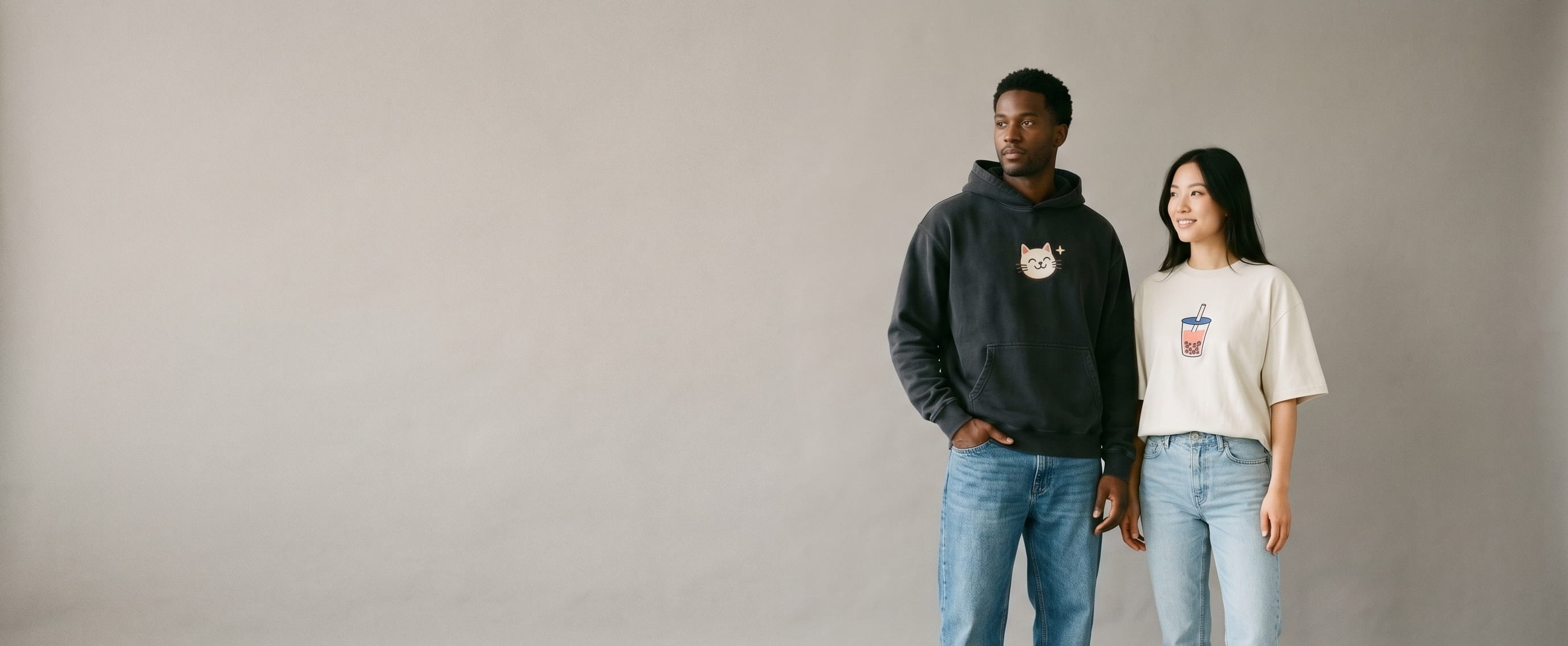 Two streetwear-capsule lifestyle models wearing decorated DTLA Print house pieces — a cartoon-cat mascot chest embroidery on the man's washed-black oversized fleece hoodie and a boba bubble-tea cup heat-press transfer on the woman's bone oversized cotton tee