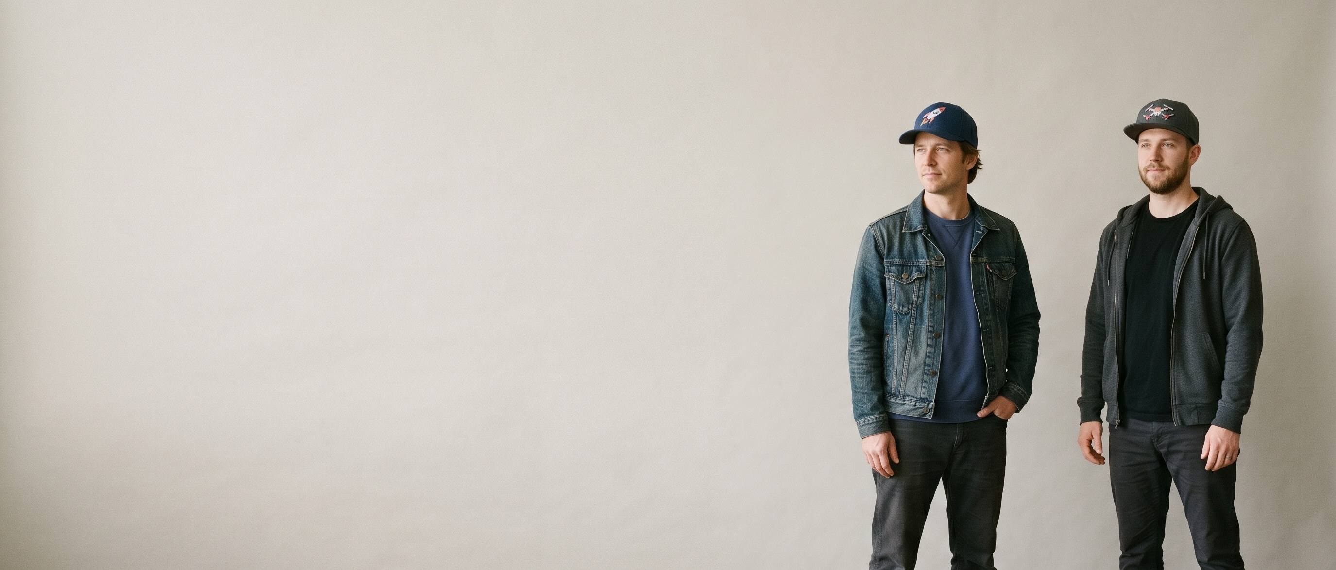 Two performance-lifestyle models wearing decorated Flexfit caps — a front-crown rocket embroidery on the first man's navy Flexfit fitted cap and a front-crown drone 3D puff on the second man's charcoal One Ten snapback