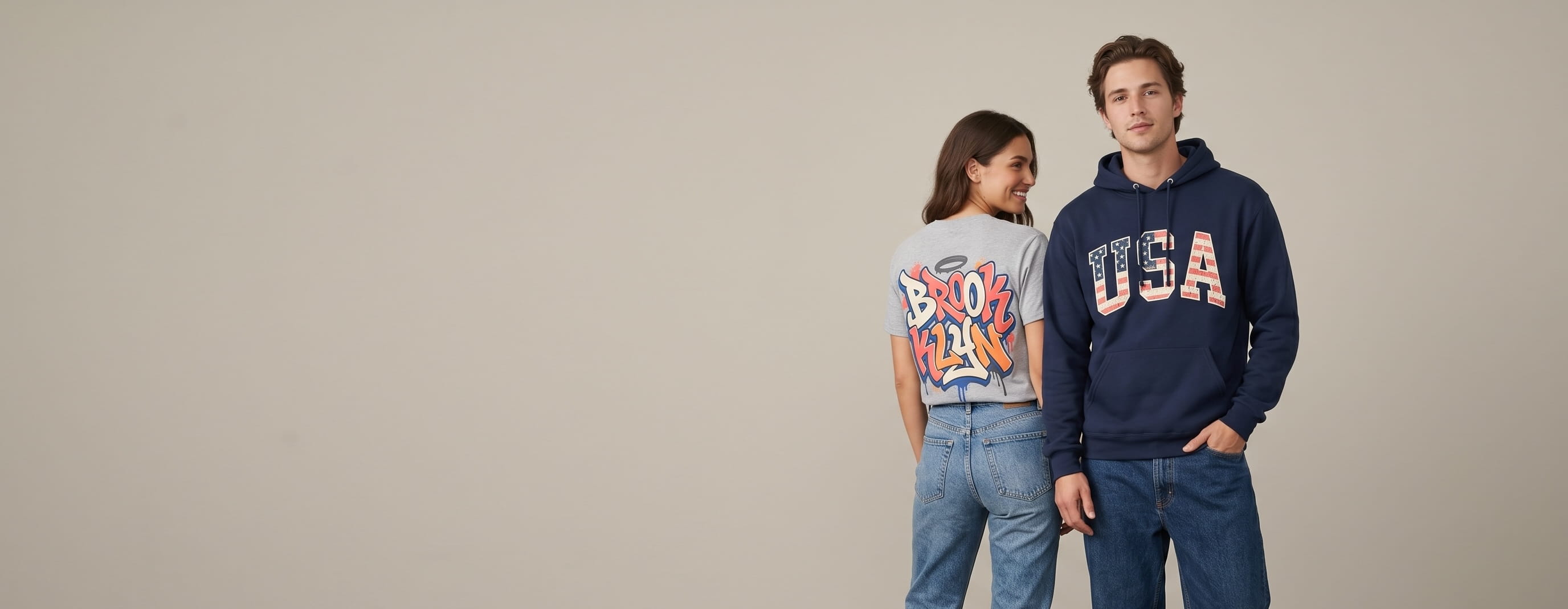 Two everyday-lifestyle models wearing decorated Fruit of the Loom — a large full-front USA Americana wordmark screen print on the man's navy Sofspun pullover hoodie, with the woman turned to show a large full-back Brooklyn graffiti heat-press transfer on her heather-grey HD Cotton crewneck tee