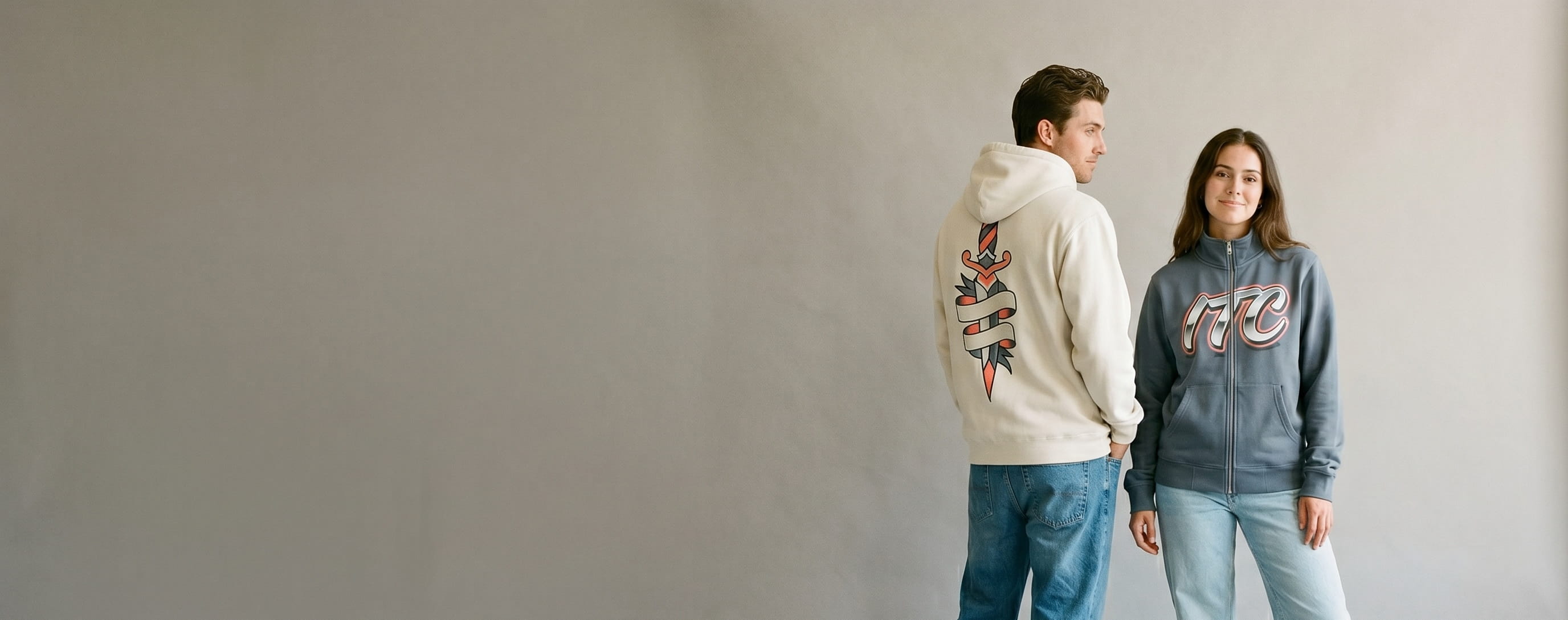 Two streetwear lifestyle models wearing decorated Independent Trading Co. — full-back traditional tattoo dagger plastisol screen print on the man's bone heavyweight pullover hoodie and an airbrushed-chrome wordmark full-color DTG on the woman's slate French Terry full-zip hoodie