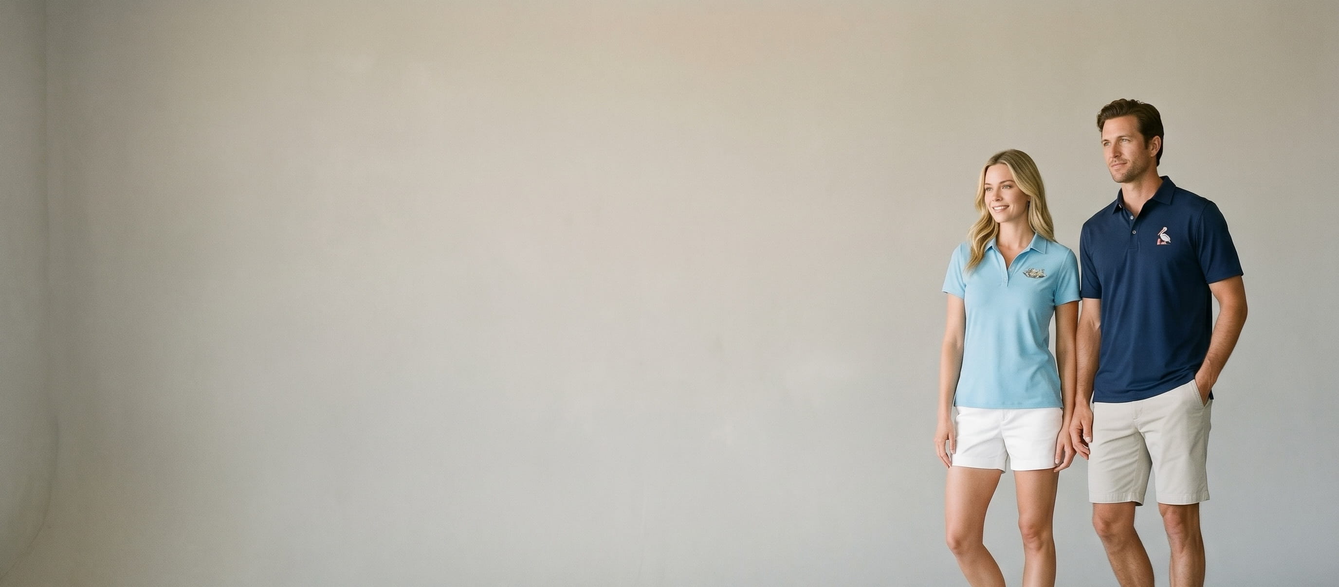 Two coastal-prep lifestyle models wearing decorated johnnie-O polos — a pelican left-chest embroidery on the man's navy prep performance polo and a sea-otter left-chest heat-press transfer on the woman's surf-blue Sadie performance polo
