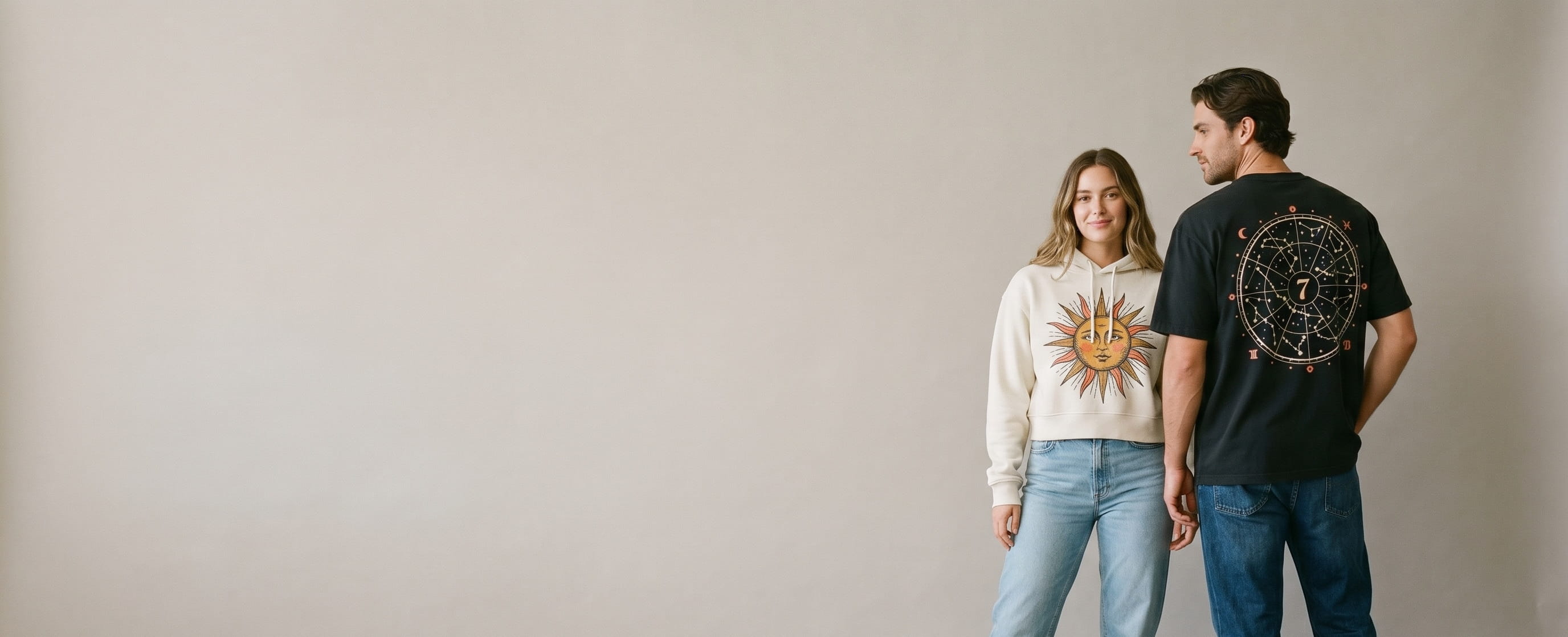 Two premium-streetwear models wearing decorated Lane Seven pieces — a large full-back zodiac constellation star-map plastisol screen print on the man's vintage-black heavyweight cotton tee, with the woman facing front in a bone ladies cropped fleece hoodie showing a celestial sun-face full-color DTG graphic