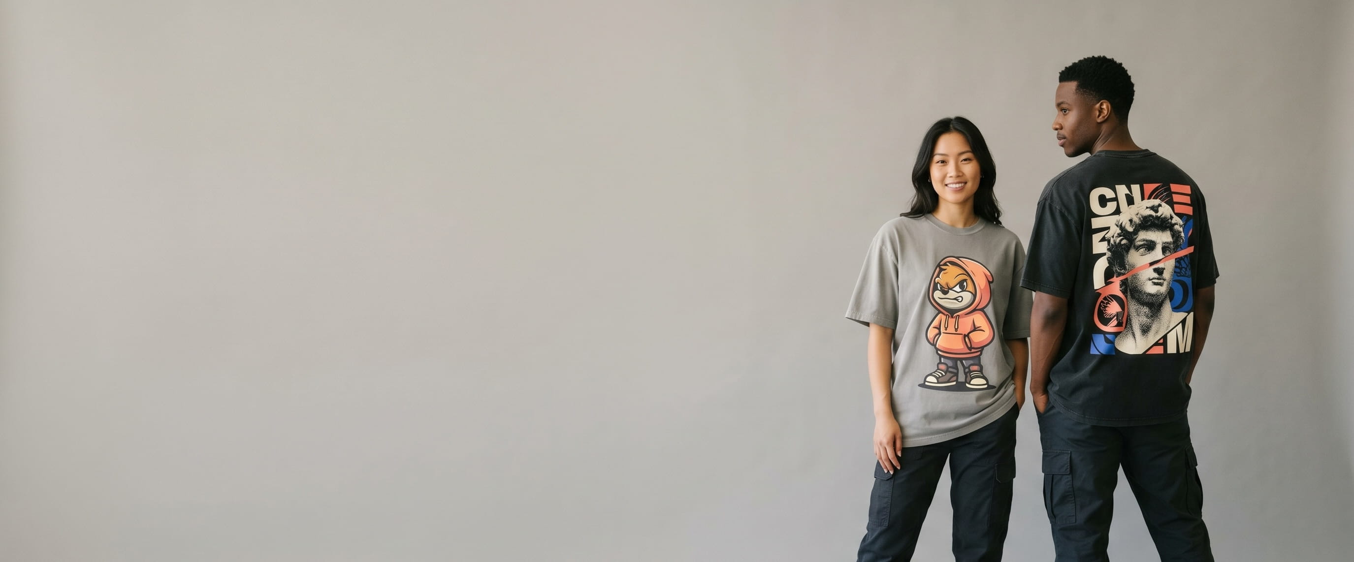 Two streetwear lifestyle models wearing decorated Los Angeles Apparel — a full-back googly-eyeball plastisol screen print on the man's washed-black garment-dye heavyweight tee and a full-chest flame-character full-color DTG on the woman's cement-grey garment-dye heavyweight tee