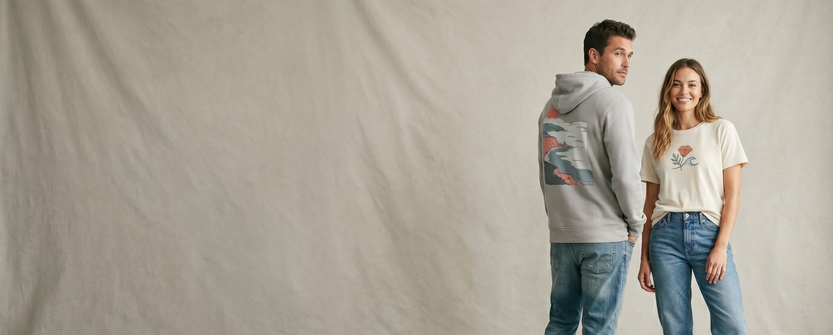Two soft-casual California lifestyle models wearing decorated Marine Layer — a full-back coastal fog-bank soft-hand screen print on the man's fog-grey Sunset pullover hoodie and a minimal serif wordmark full-color DTG on the woman's natural-cream Signature super-soft tee