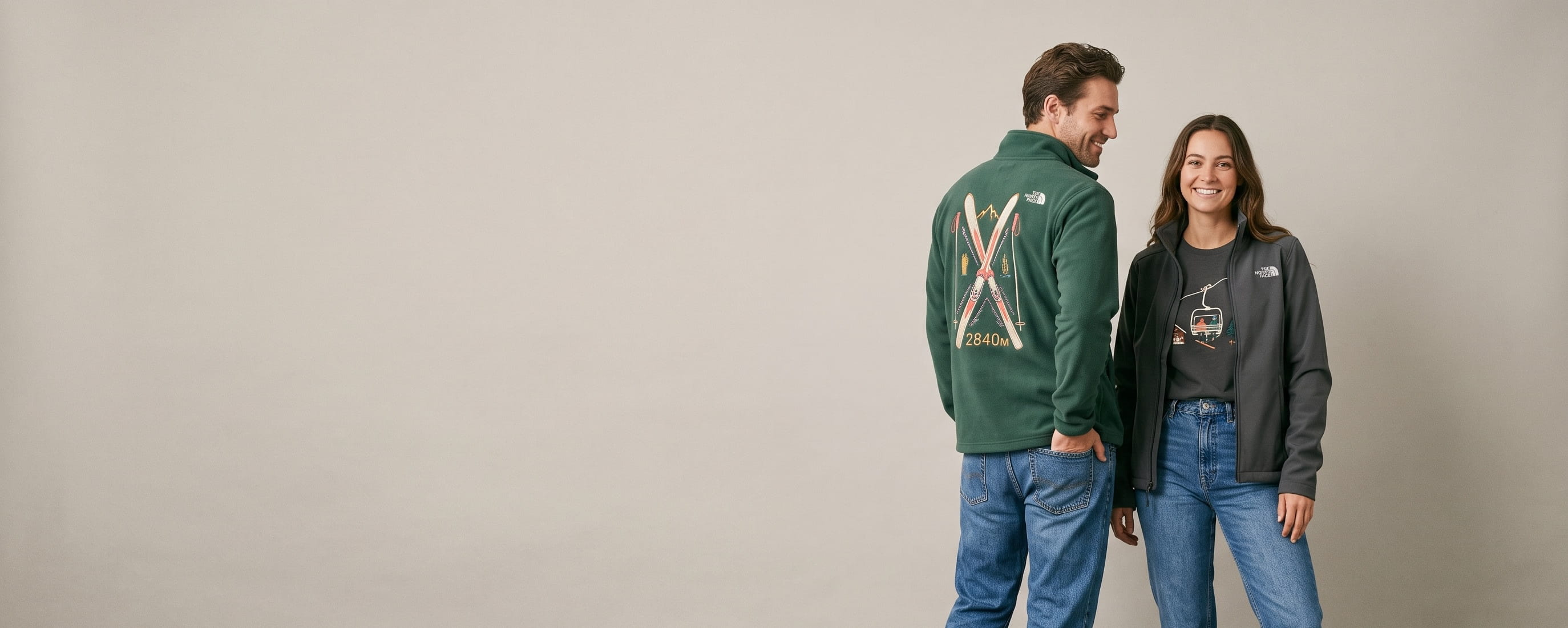 Two outdoor-lifestyle models wearing decorated North Face outerwear — a large full-back crossed vintage-skis embroidery on the man's pine-green full-zip fleece jacket, with the woman facing front in an asphalt-grey Apex soft shell showing a chairlift chest embroidery