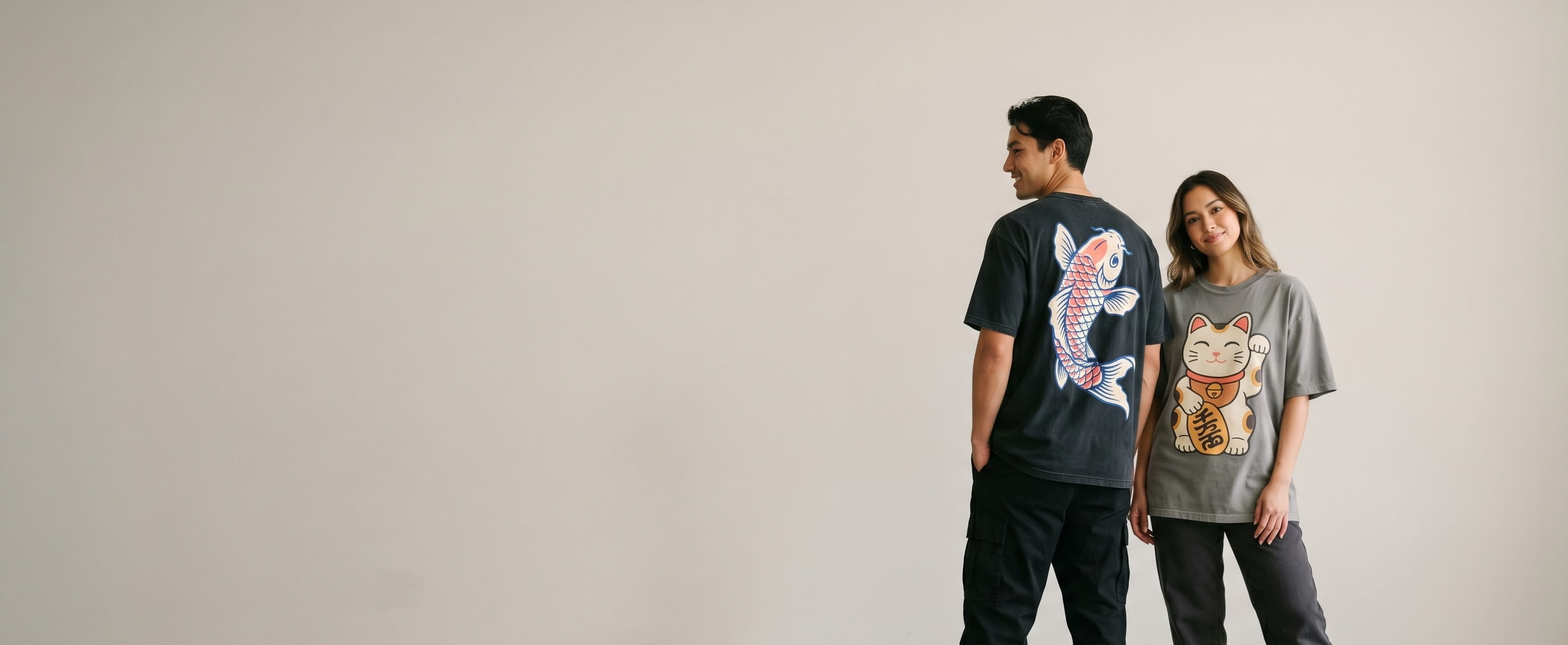 Two streetwear lifestyle models wearing decorated Shaka Wear — a full-back koi plastisol screen print on the man's washed-black heavyweight tee and a full-chest lucky-cat-mascot full-color DTG on the woman's cement-grey garment-dyed drop-shoulder tee