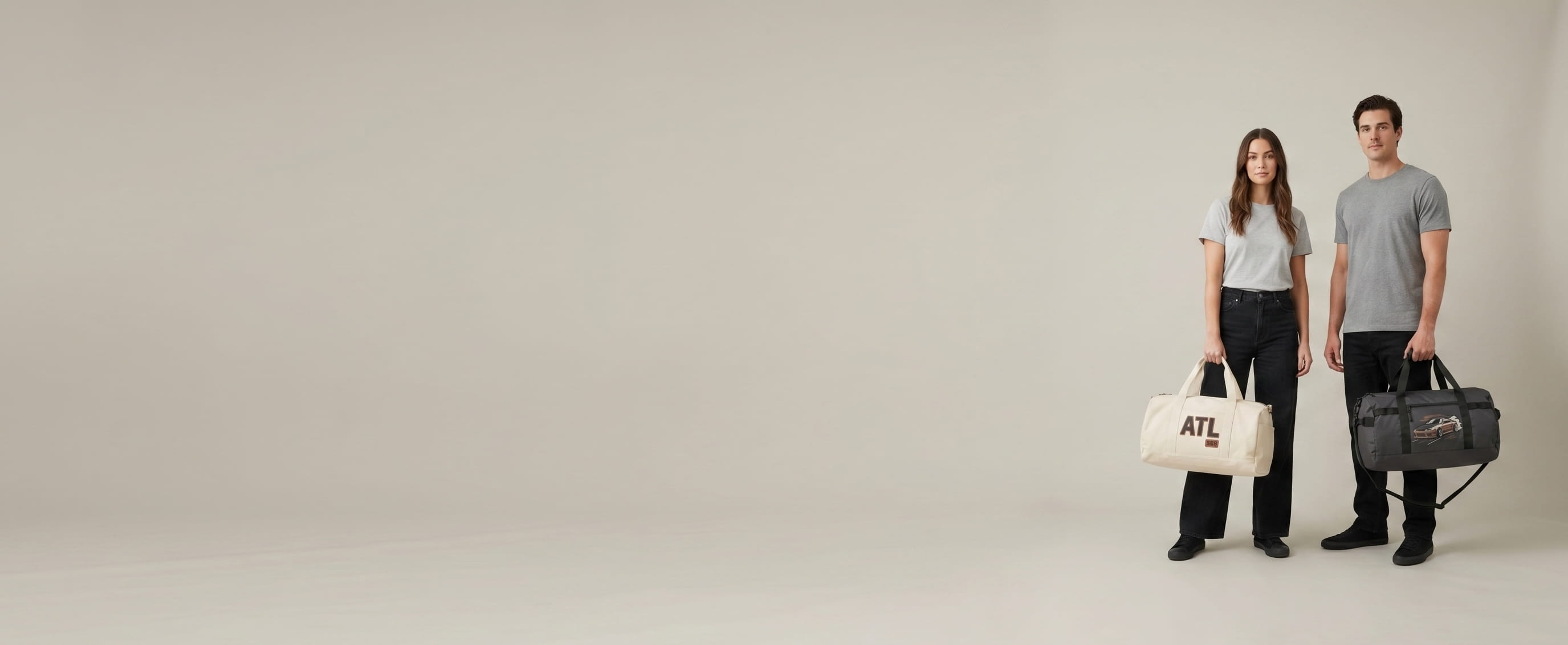 One woman + one man lifestyle models on a warm-grey studio backdrop, each carrying one decorated specialty bag held by the top handle with the front panel facing camera — one cream cotton canvas duffel with a woven-patch front-panel composition + one charcoal-gray polyester ripstop duffel with a hand-painted front-panel composition