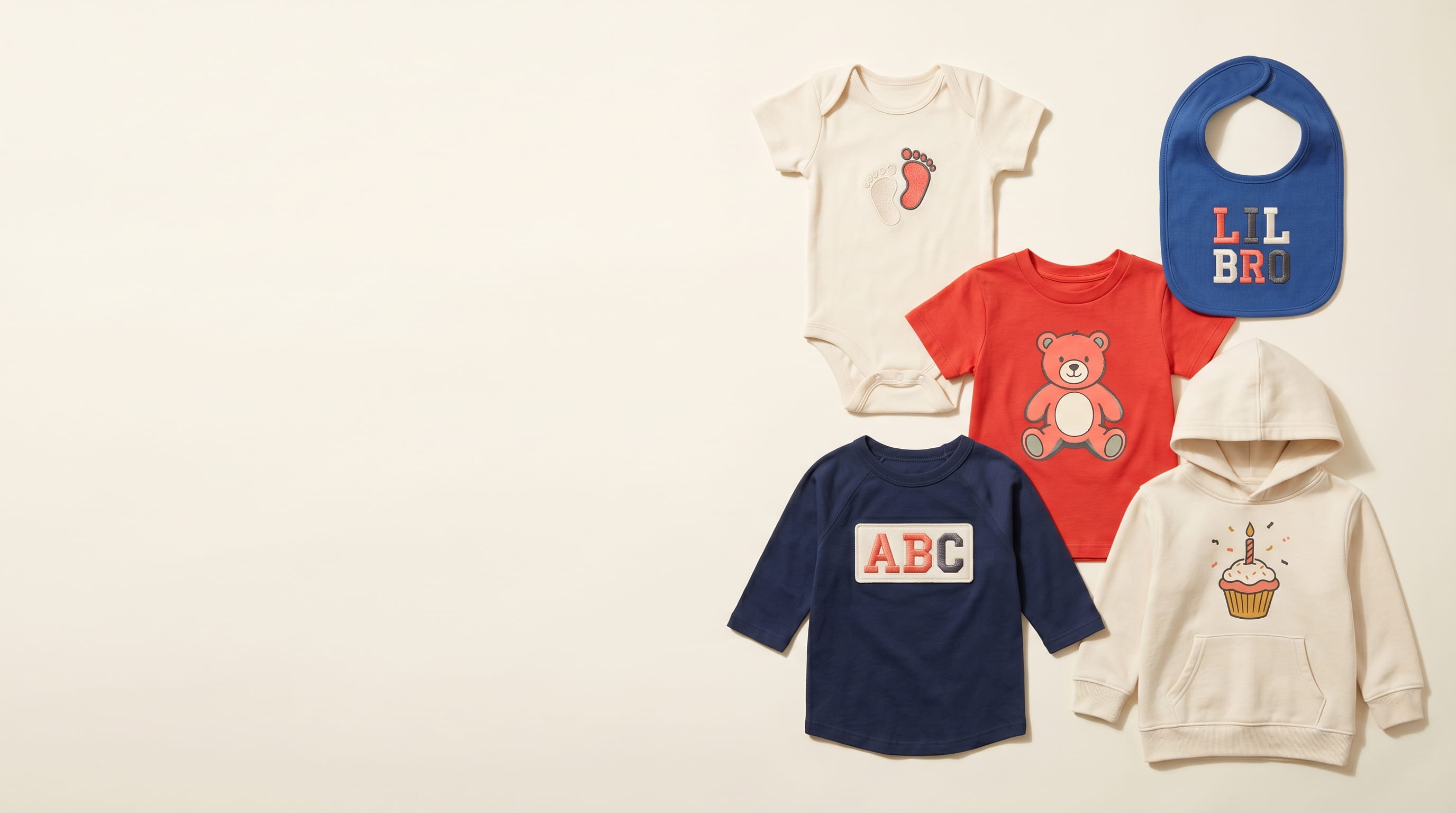 Five decorated toddler + infant apparel pieces arranged magazine-style flat-lay on a warm-white studio backdrop — cream baby-rib onesie with heritage baby footprint emblem, cobalt baby-rib bib with heritage sibling badge, scarlet fine-jersey toddler tee with full-color DTG baby-bear emblem, navy poly-cotton toddler raglan with heritage embroidered ABC alphabet sampler patch, cream fleece toddler hoodie with heat-press celebration cupcake emblem