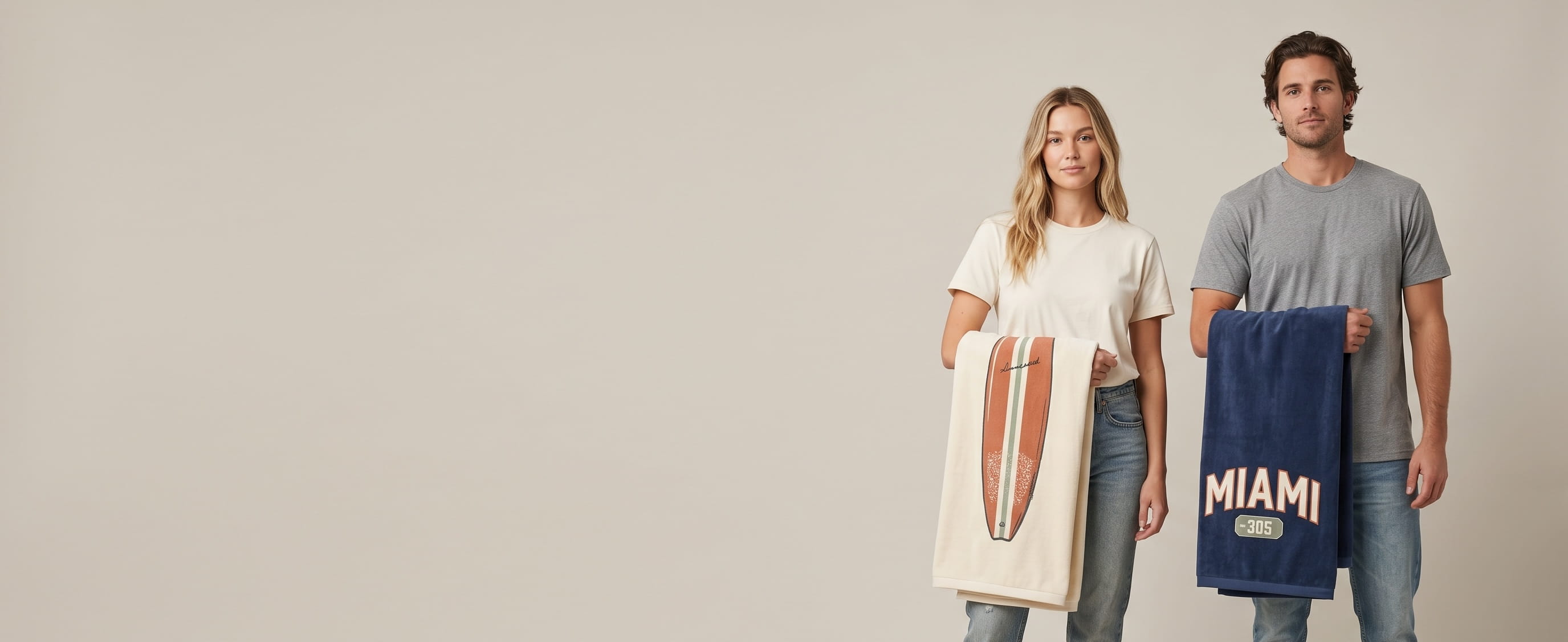 One woman + one man lifestyle models on a warm-grey studio backdrop, each holding one decorated folded towel over the forearm with the decorated corner facing camera — one cream velour beach towel with a hand-painted vintage resort screen-print composition + one navy velour beach towel with a heritage woven city-code patch composition