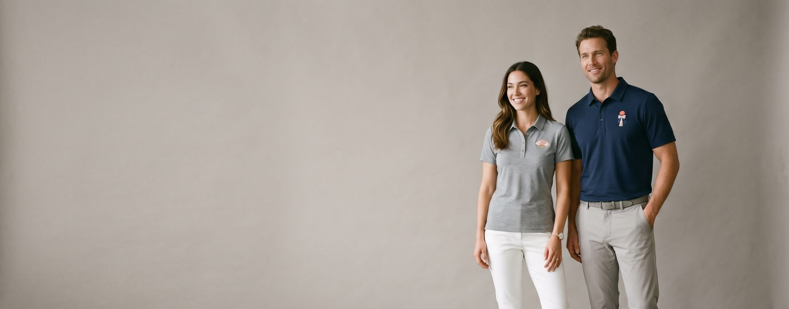 Two premium golf-lifestyle models wearing decorated TravisMathew — a left-chest centurion-helmet chain-stitch on the man's navy Coto performance polo and a left-chest cherub embroidery on the woman's heather-grey Oceanside performance polo