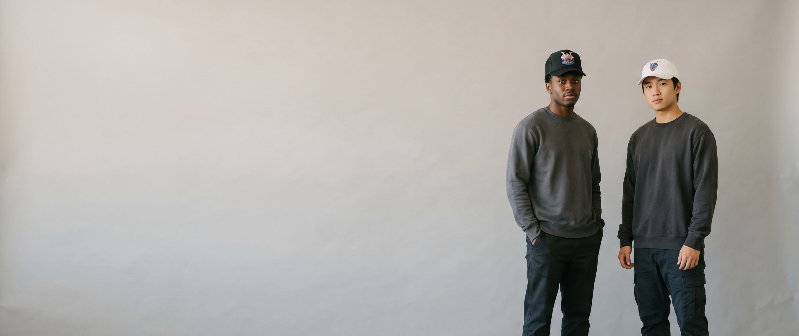 Two streetwear lifestyle models wearing decorated Yupoong caps — a front-crown cyber-mech-samurai-bust 3D puff on the first man's black foam-front trucker and a front-crown hannya-mask embroidery on the second man's white dad hat