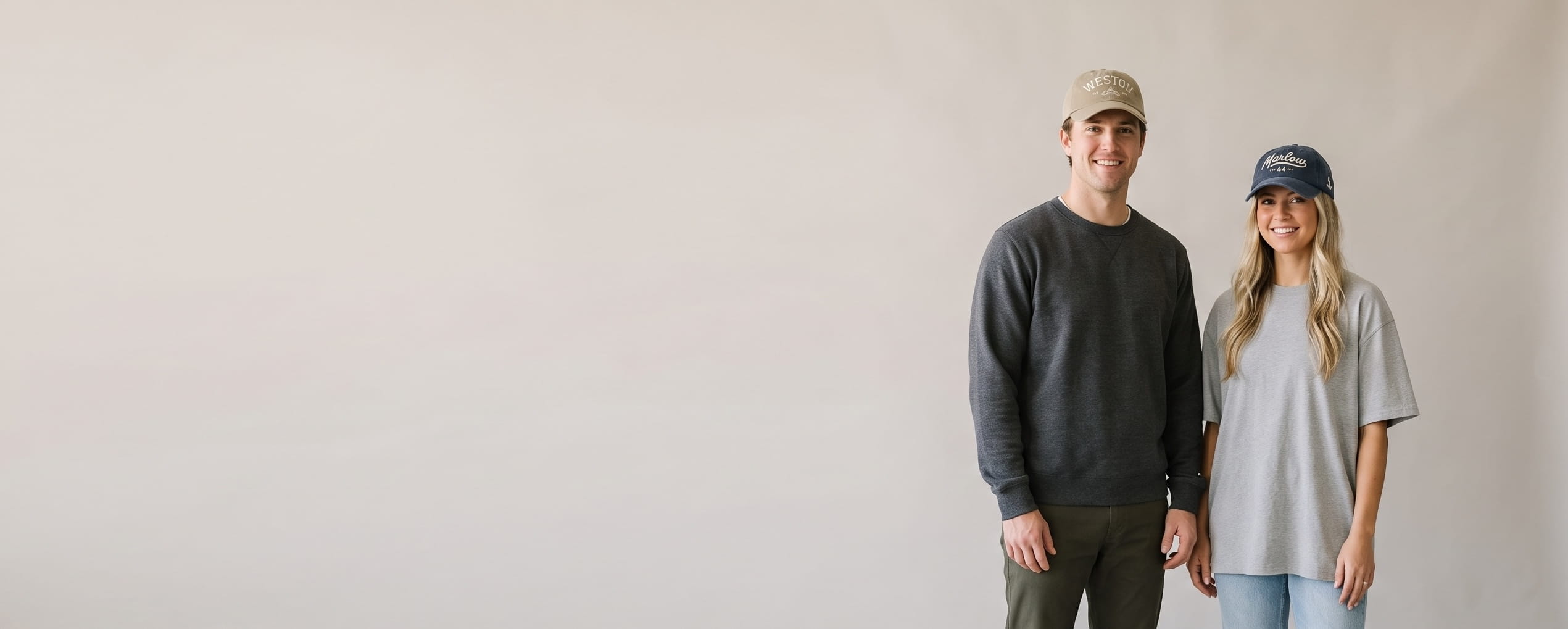 Two lifestyle models in decorated custom dad hats — man in a washed khaki unstructured dad cap with a tonal flat-embroidered wordmark + woman in a pigment-dyed navy dad cap with a chain-stitch script and side-panel emblem