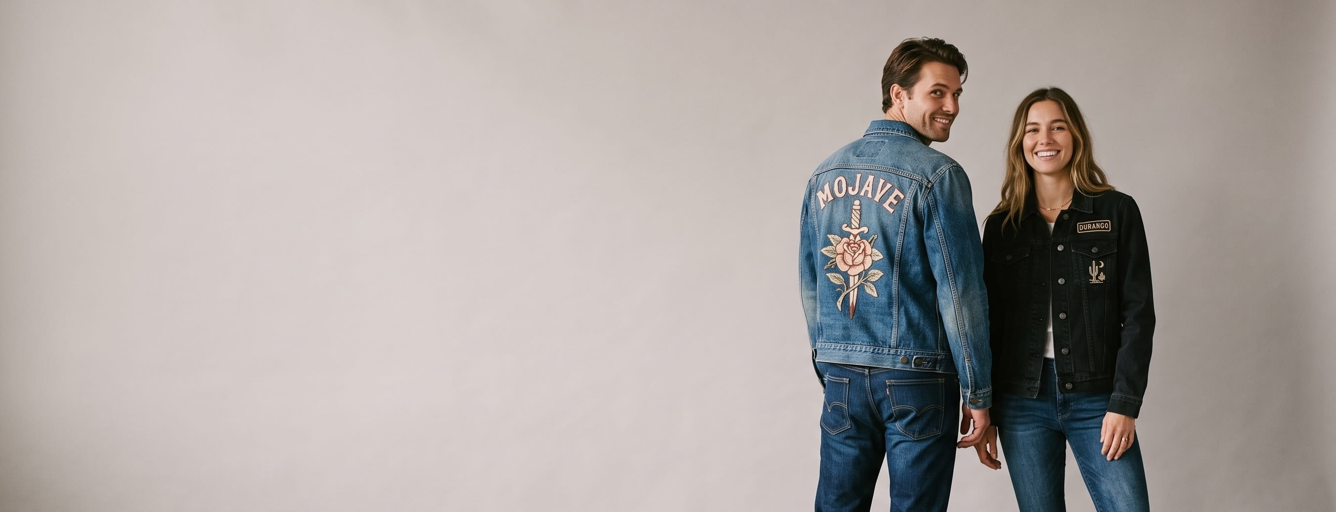 Two lifestyle models in decorated custom denim jackets — man in a mid-wash trucker denim jacket with a full-back chain-stitch graphic + woman in a black denim jacket with an embroidered back-panel emblem and chest patches