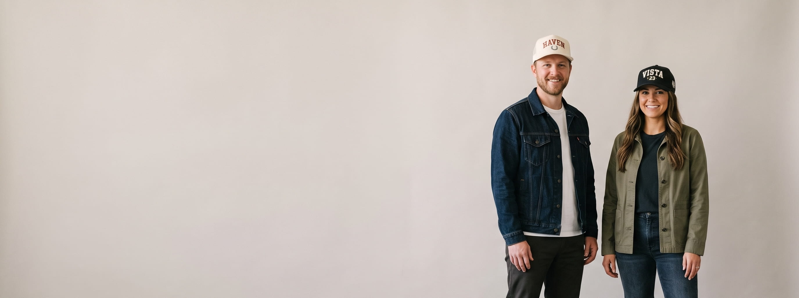 Two lifestyle models in decorated custom hats — man in a natural cotton-twill trucker cap with a flat-embroidered wordmark + woman in a charcoal structured cap with a 3D puff monogram and side-panel chain-stitch