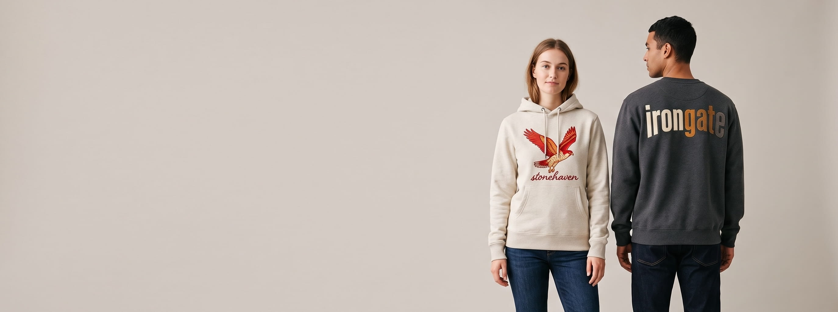 Two lifestyle models in decorated custom hoodies and sweaters — woman in an oatmeal cotton-poly fleece pullover hoodie with a full-chest embroidered graphic + man in an anthracite heavyweight brushed-fleece crewneck with a full-back embroidered wordmark