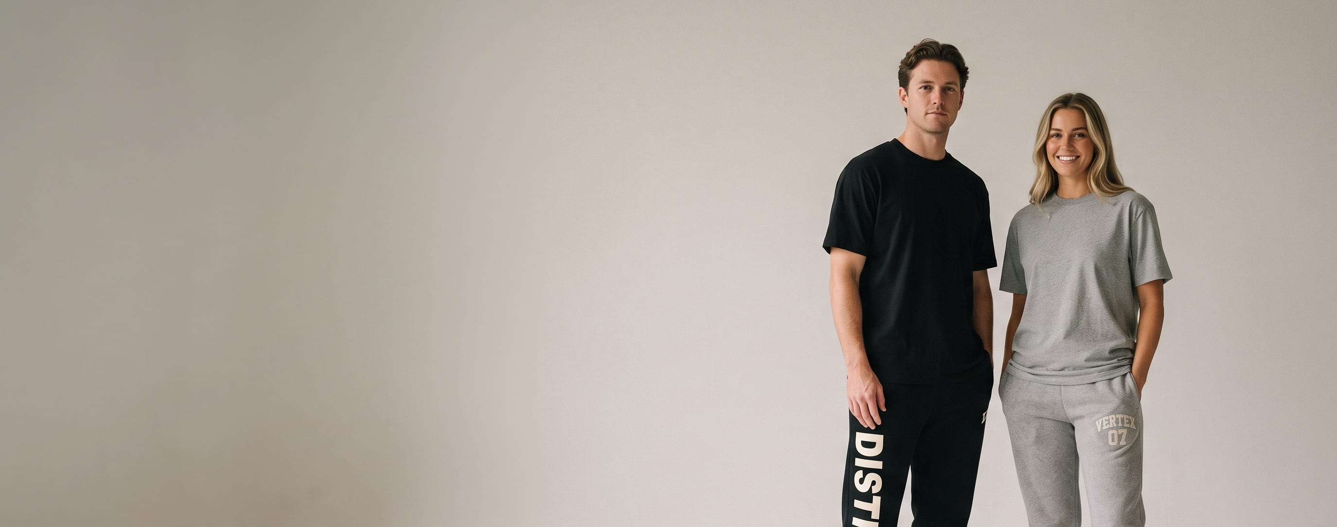 Two lifestyle models in decorated custom joggers — man in heavyweight fleece cuffed joggers with a vertical side-seam wordmark down the leg + woman in sueded fleece joggers with a front-thigh embroidered emblem