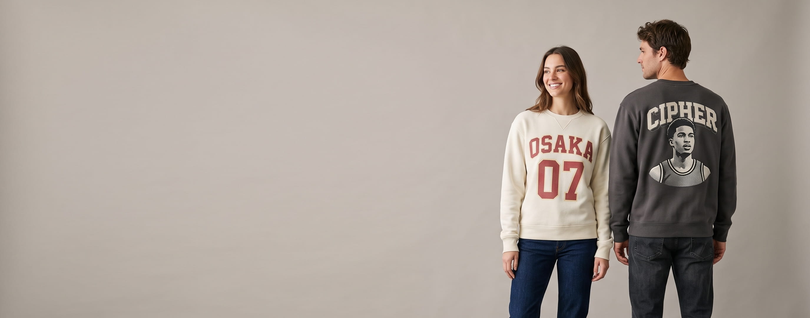 Two lifestyle models in decorated custom pullover sweatshirts — woman in an ivory cotton-poly fleece crewneck with a full-chest varsity graphic + man in an anthracite heavyweight brushed-fleece crewneck with a full-back embroidered graphic