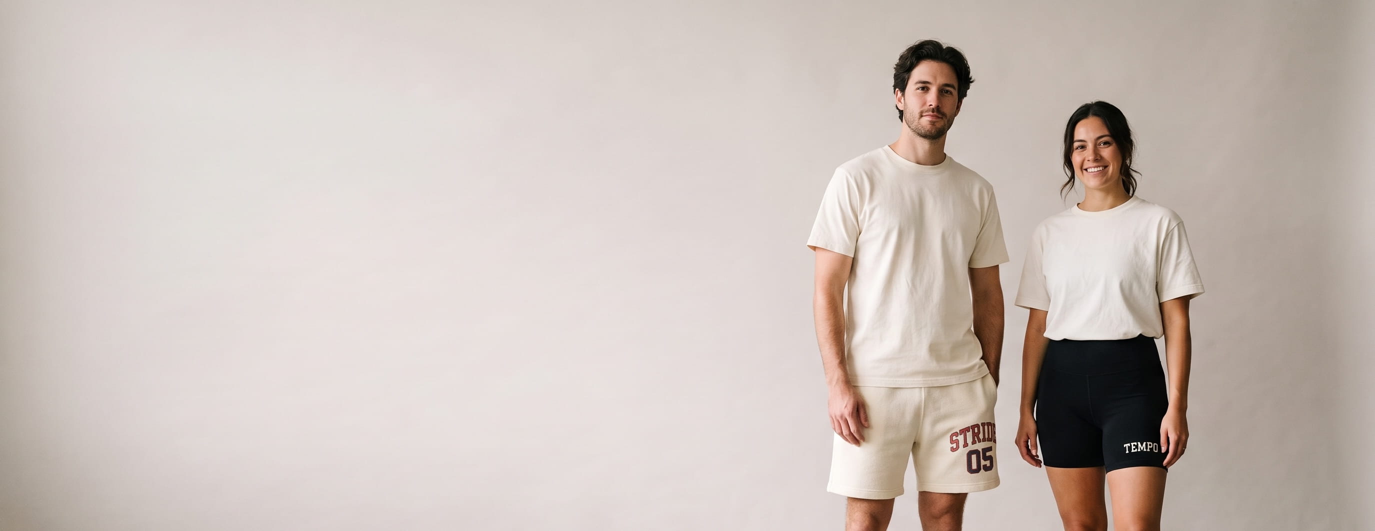 Two lifestyle models in decorated custom shorts — man in heavyweight fleece sweat shorts with a screen-printed hip logo + woman in high-waist biker shorts with an embroidered leg wordmark