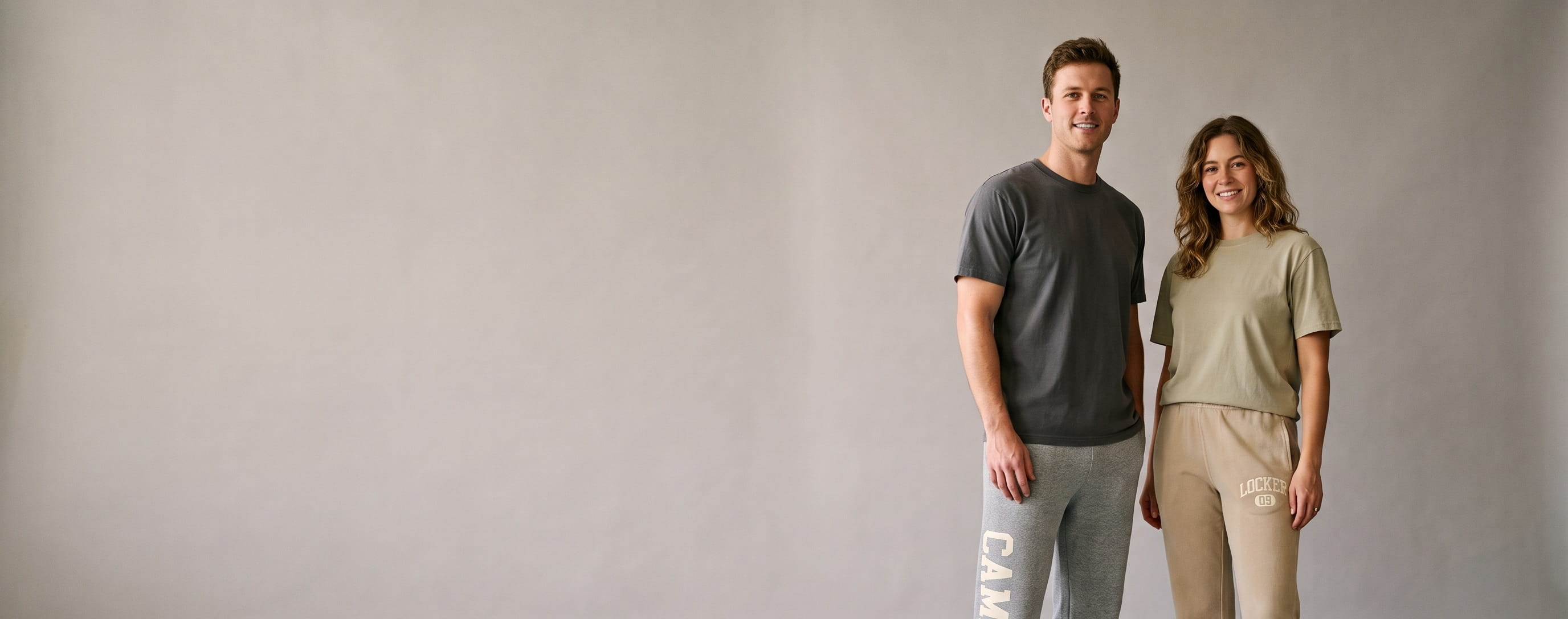 Two lifestyle models in decorated custom sweatpants — man in heavyweight open-bottom fleece sweatpants with a vertical side-seam wordmark down the leg + woman in garment-dyed straight-leg sweatpants with a front-thigh embroidered emblem