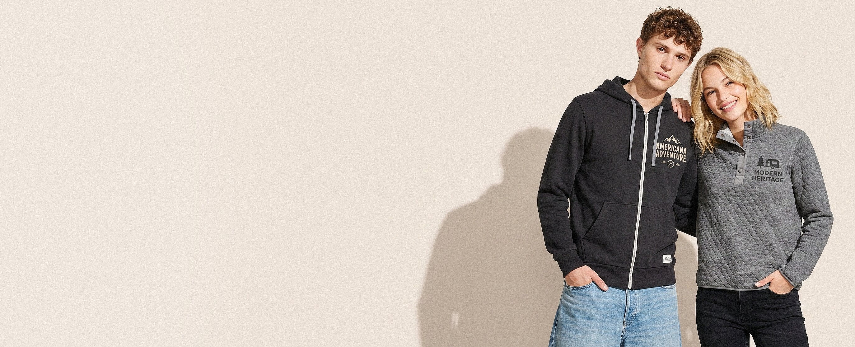 Two soft-casual California lifestyle models wearing decorated Marine Layer — a full-back coastal fog-bank soft-hand screen print on the man's fog-grey Sunset pullover hoodie and a minimal serif wordmark full-color DTG on the woman's natural-cream Signature super-soft tee
